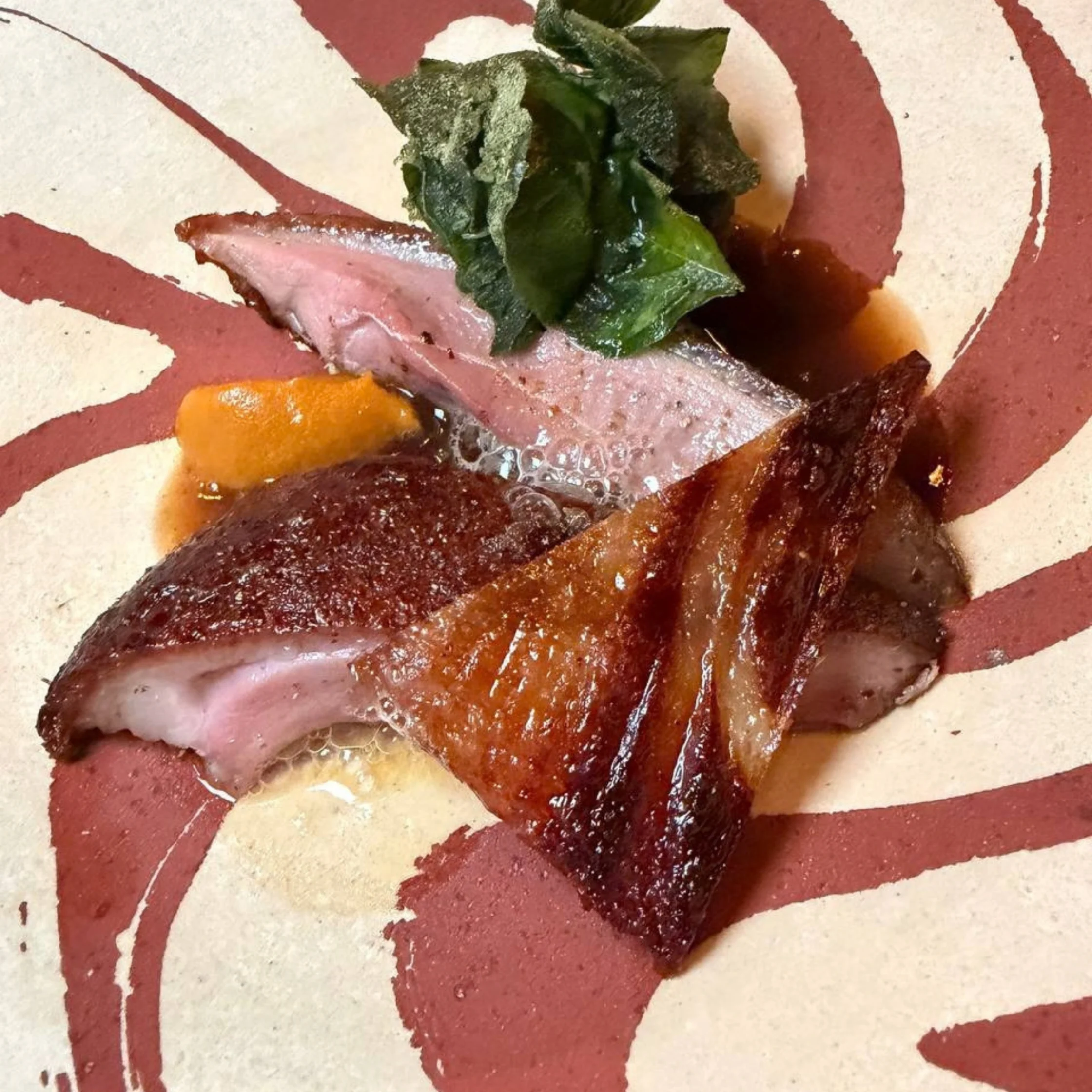 A rustic plate of glistening sliced roast meat with crisp browned edges, orange garnish, and dark leafy greens, arranged on a red-and-cream patterned plate.