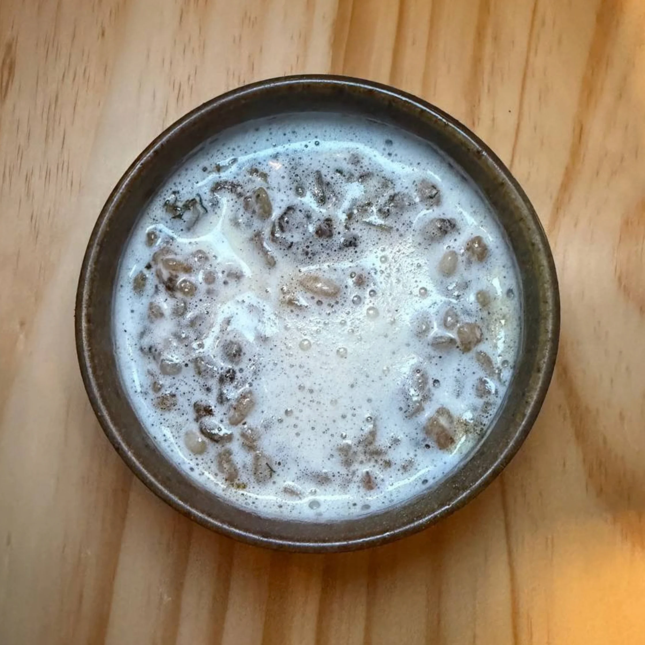 Overhead view of a dark ceramic bowl filled with creamy milk and cereal pieces on a wooden table, creating a simple, cozy breakfast scene.