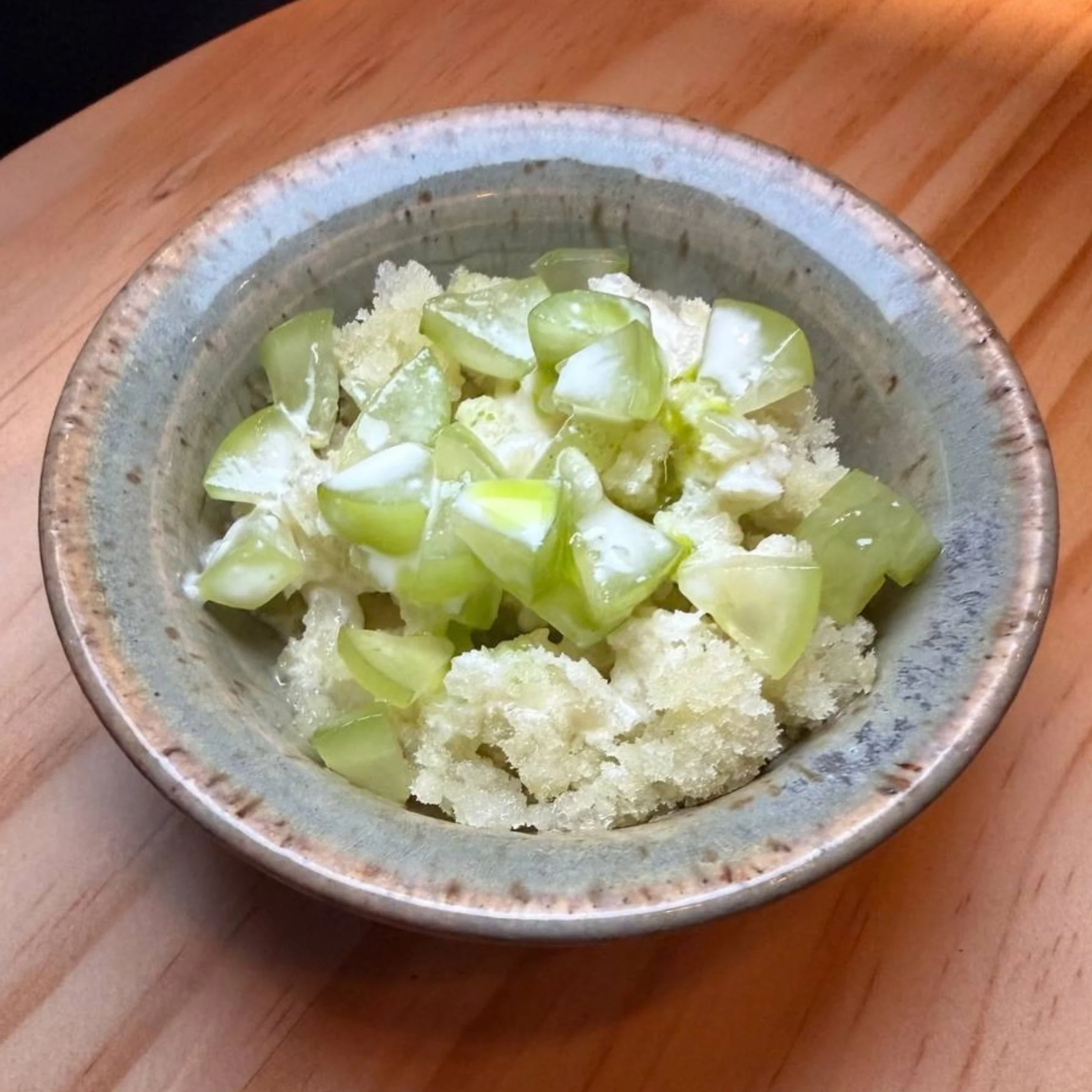 A rustic ceramic bowl holds pale green, chopped gooseberries mixed with crumbly white sugar, photographed on a wooden table in warm light.
