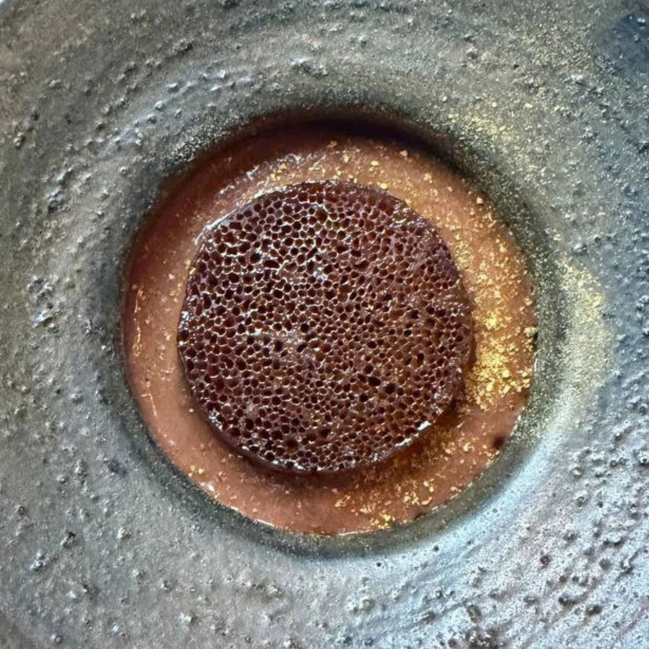 Close-up of a rusted circular metal drain or grate embedded in rough gray concrete, its honeycomb-like holes and worn bronze rim giving an aged, gritty appearance.