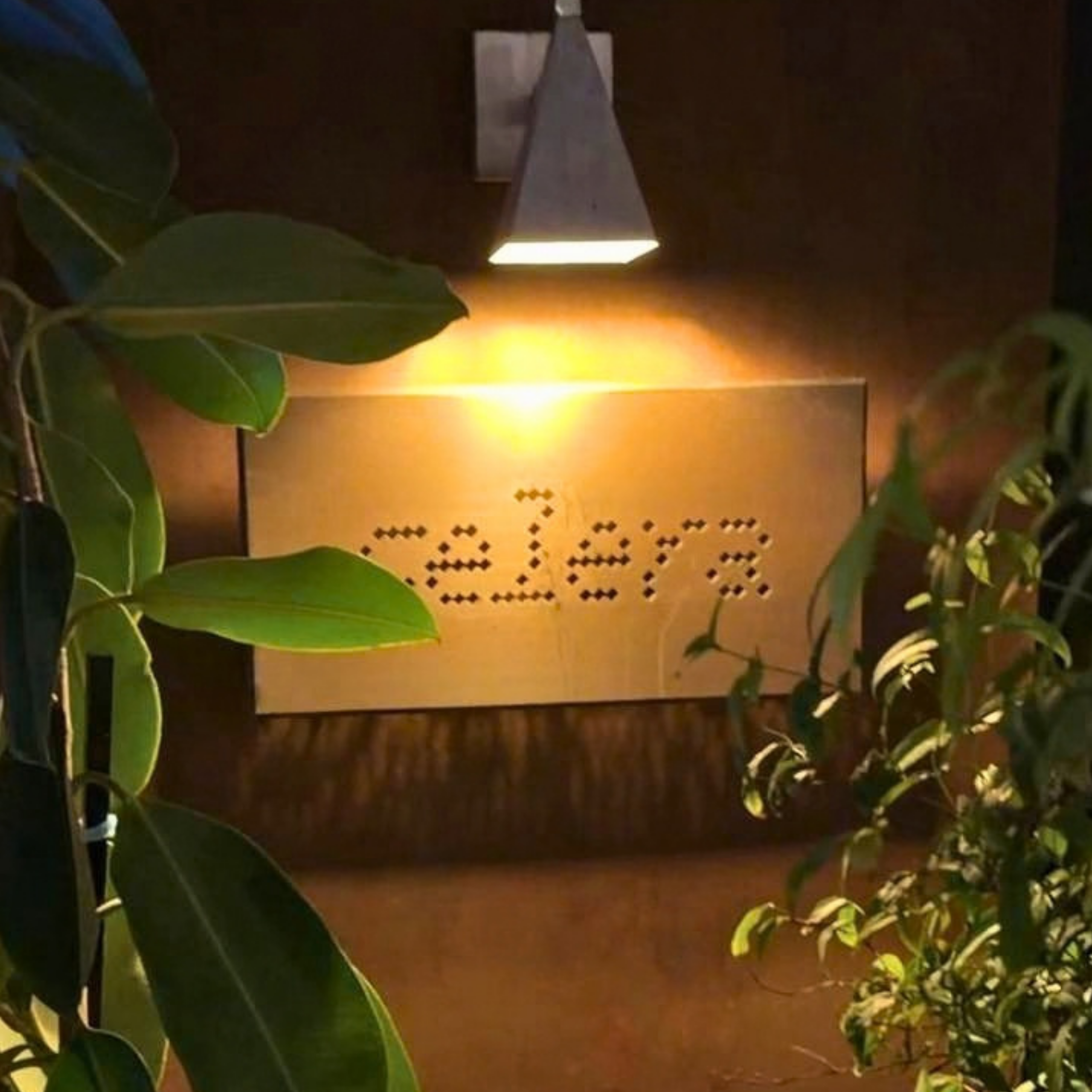 A pendant lamp casts a warm glow over a perforated sign on a dark wall, framed by lush green plants for a cozy, welcoming atmosphere.