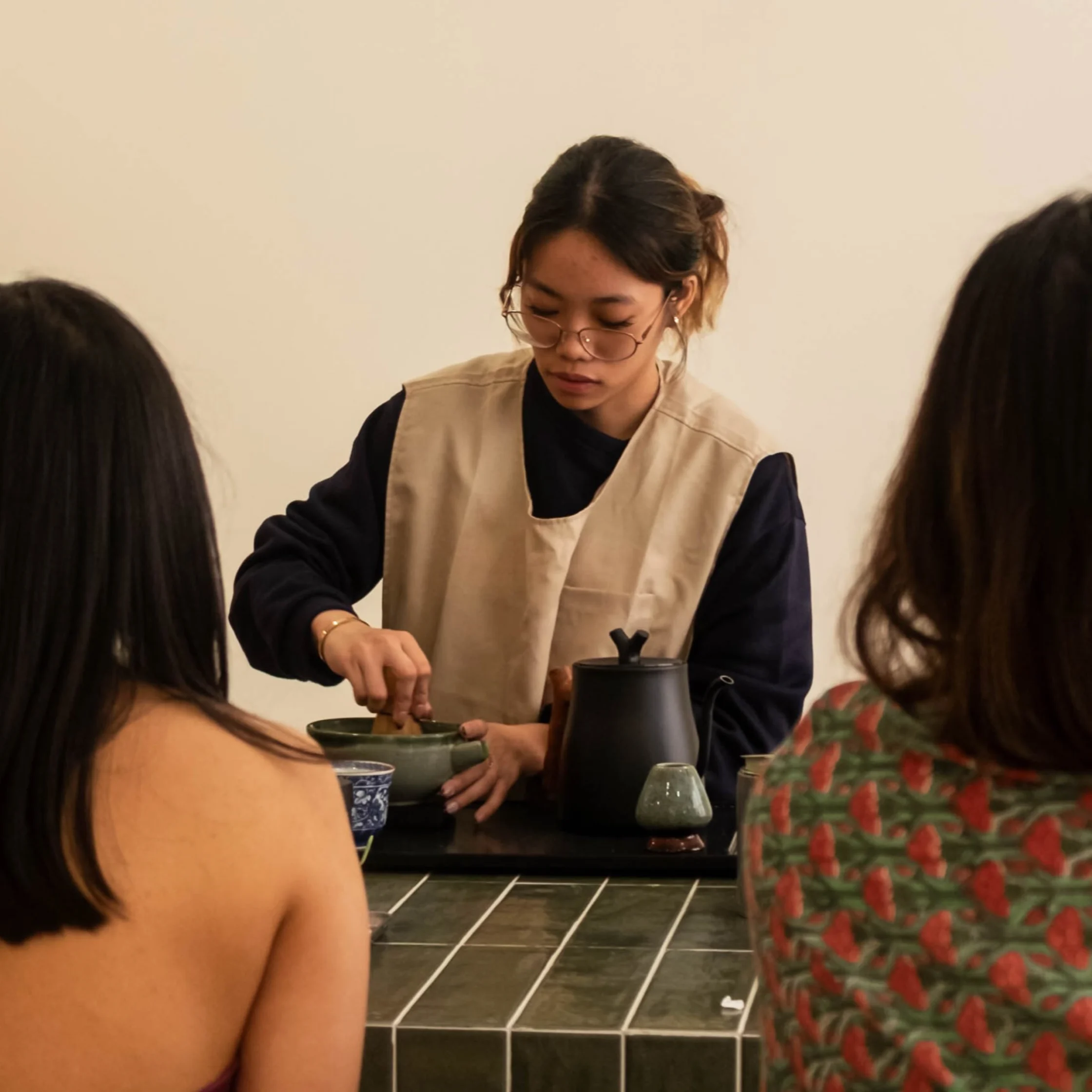 A woman in glasses and a beige apron prepares tea at a tiled counter as two people watch; the calm, warmly lit scene feels intimate and focused.