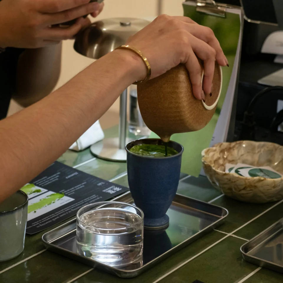 A person pours green tea from a rounded brown ceramic pot into a blue cup on a metal tray, with a water glass nearby; a calm tea service scene.