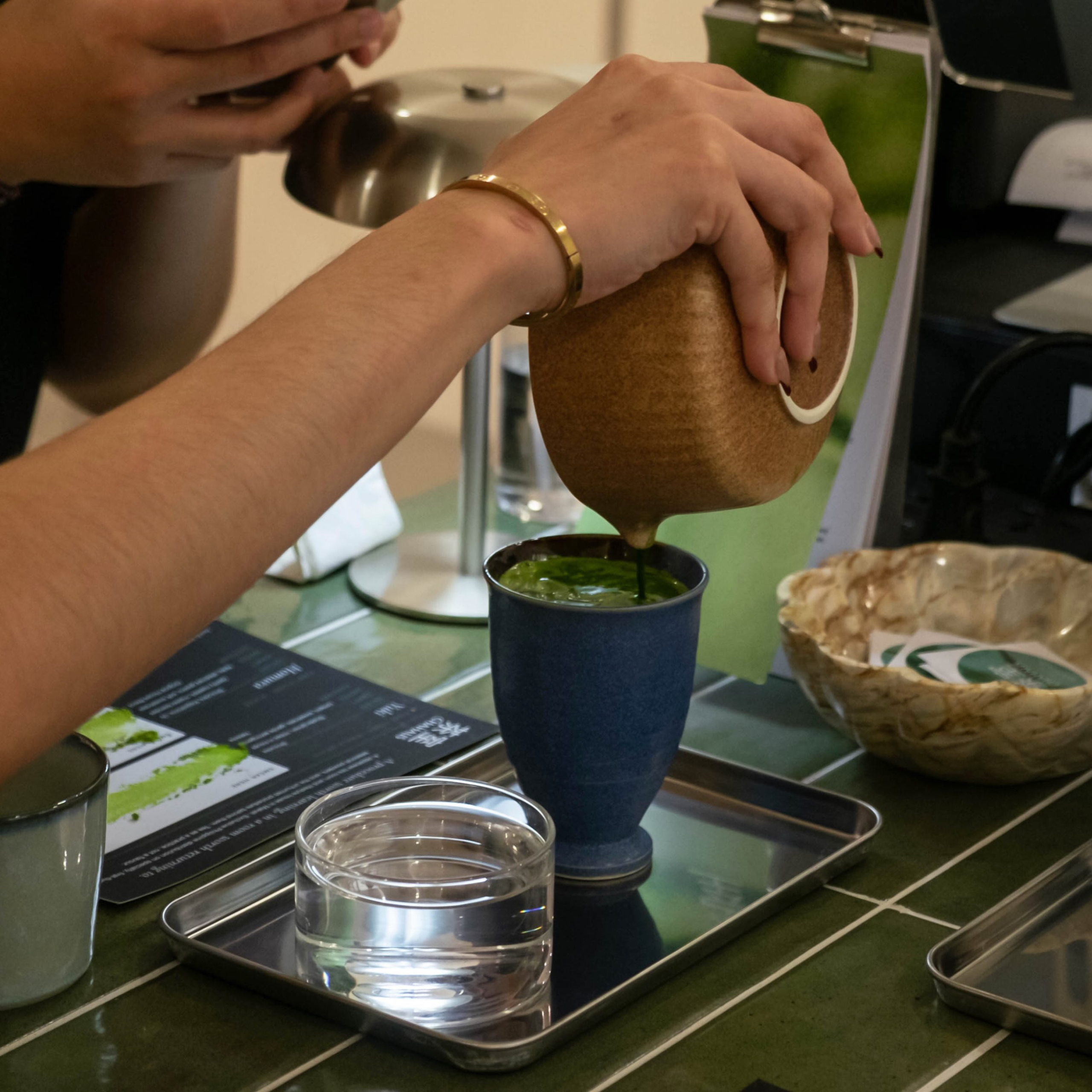 A person pours green tea from a rounded brown ceramic pot into a blue cup on a metal tray, with a water glass nearby; a calm tea service scene.