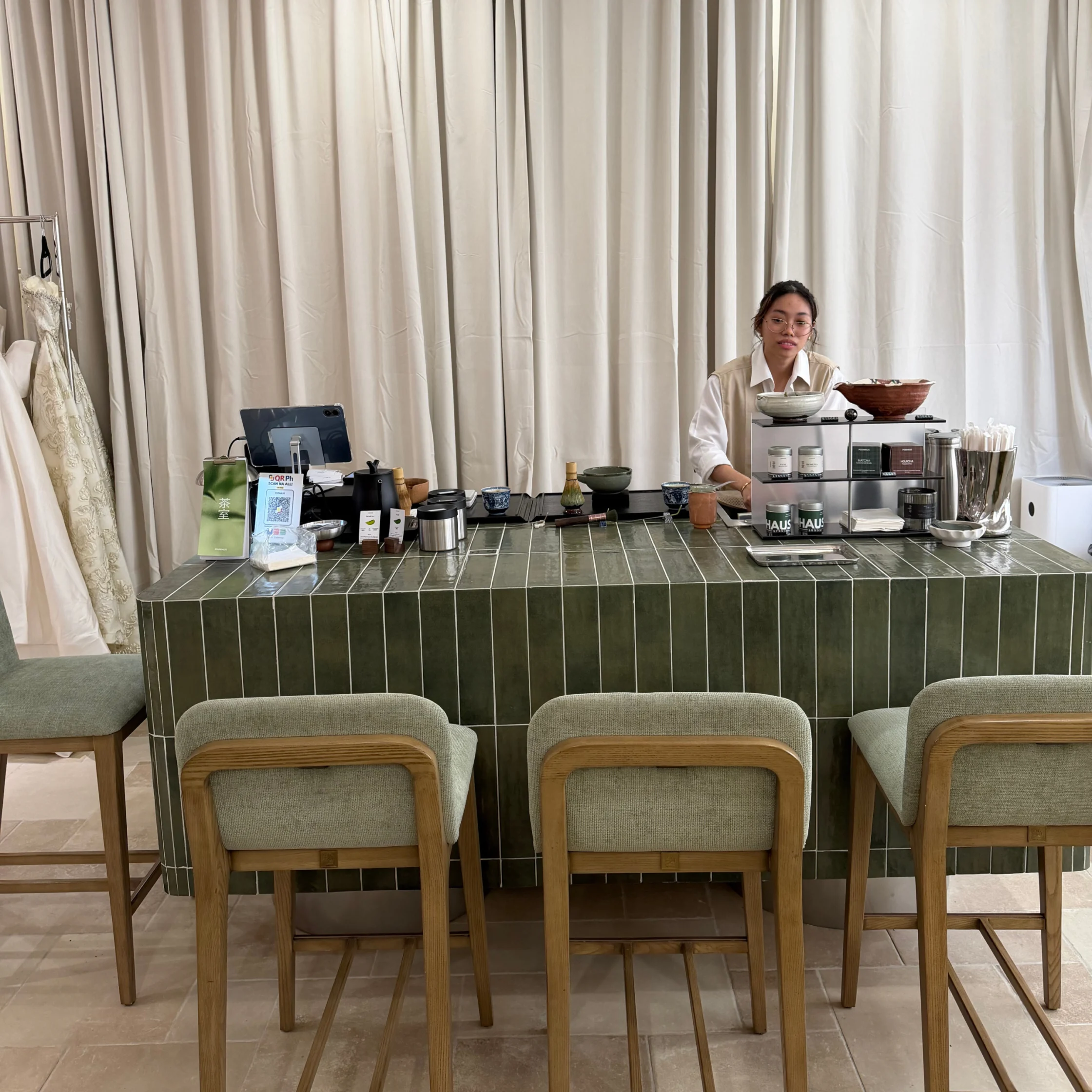 A woman stands behind a dark green tiled café counter, with coffee equipment and stools in front of cream curtains, creating a calm, minimalist atmosphere.