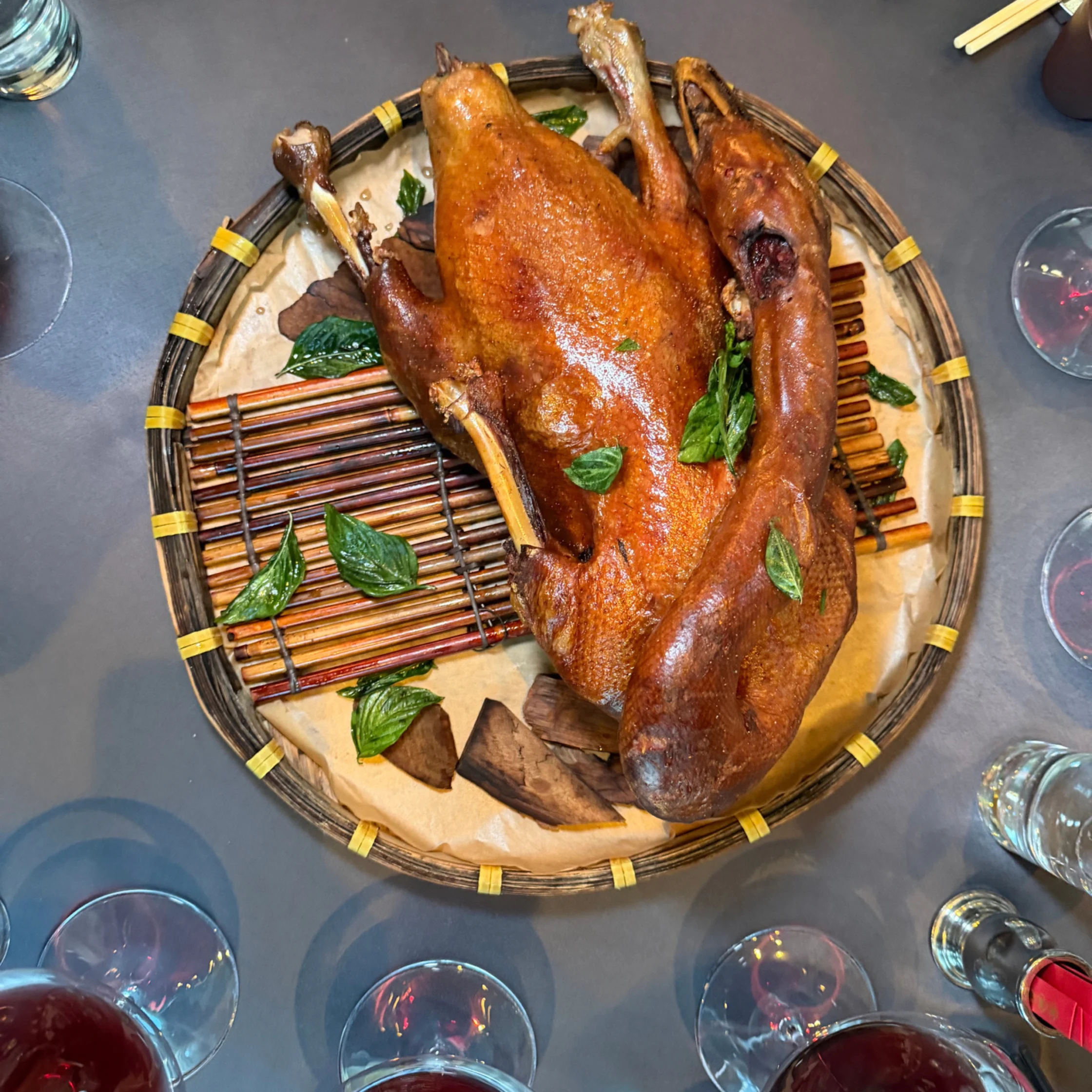 Overhead view of glossy, golden-brown roasted duck on a woven platter with green leaves, surrounded by glasses and chopsticks; a festive, inviting meal.