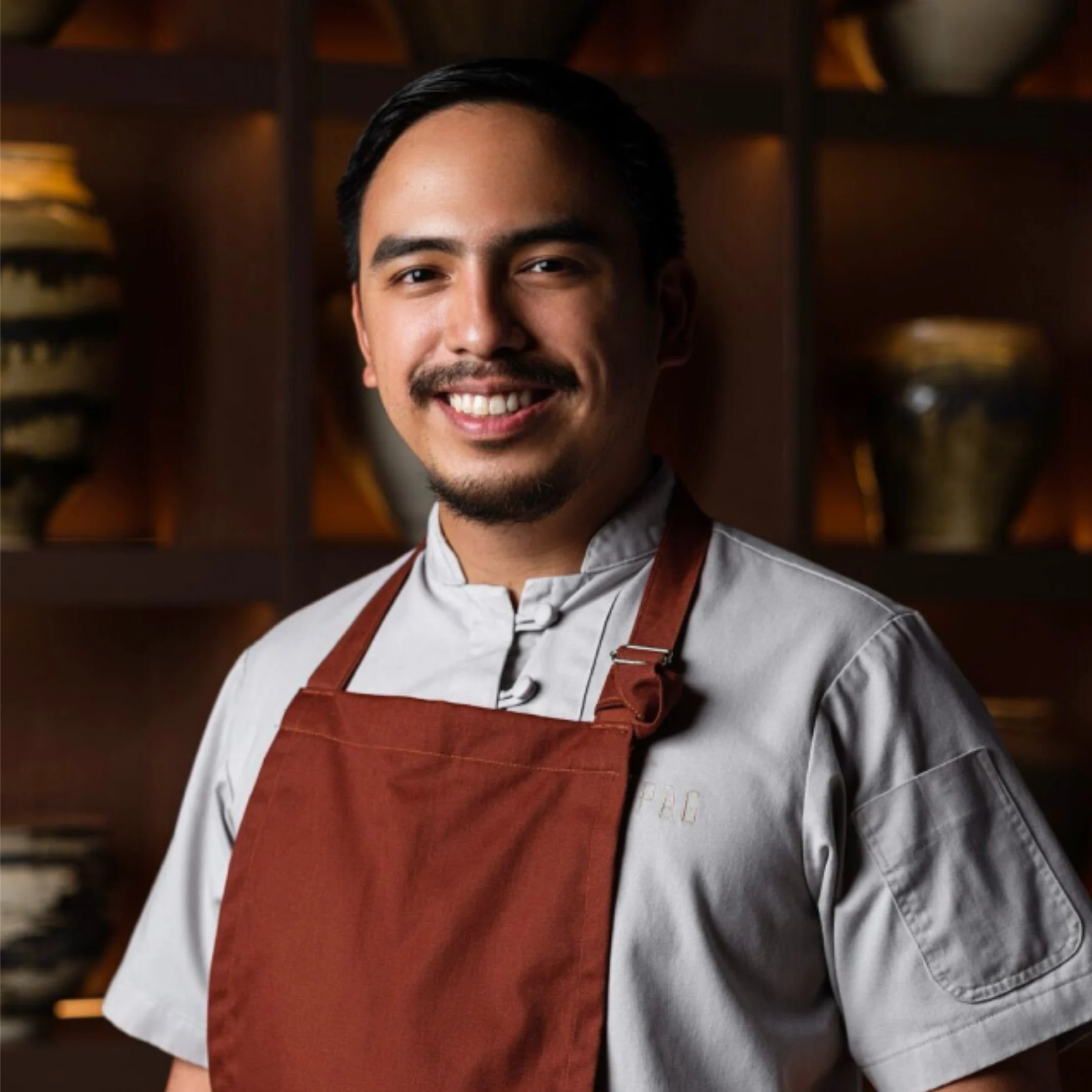 A smiling male chef in a white uniform and rust-red apron poses in a warmly lit kitchen, with shelves and cookware behind him, conveying a friendly, confident tone.