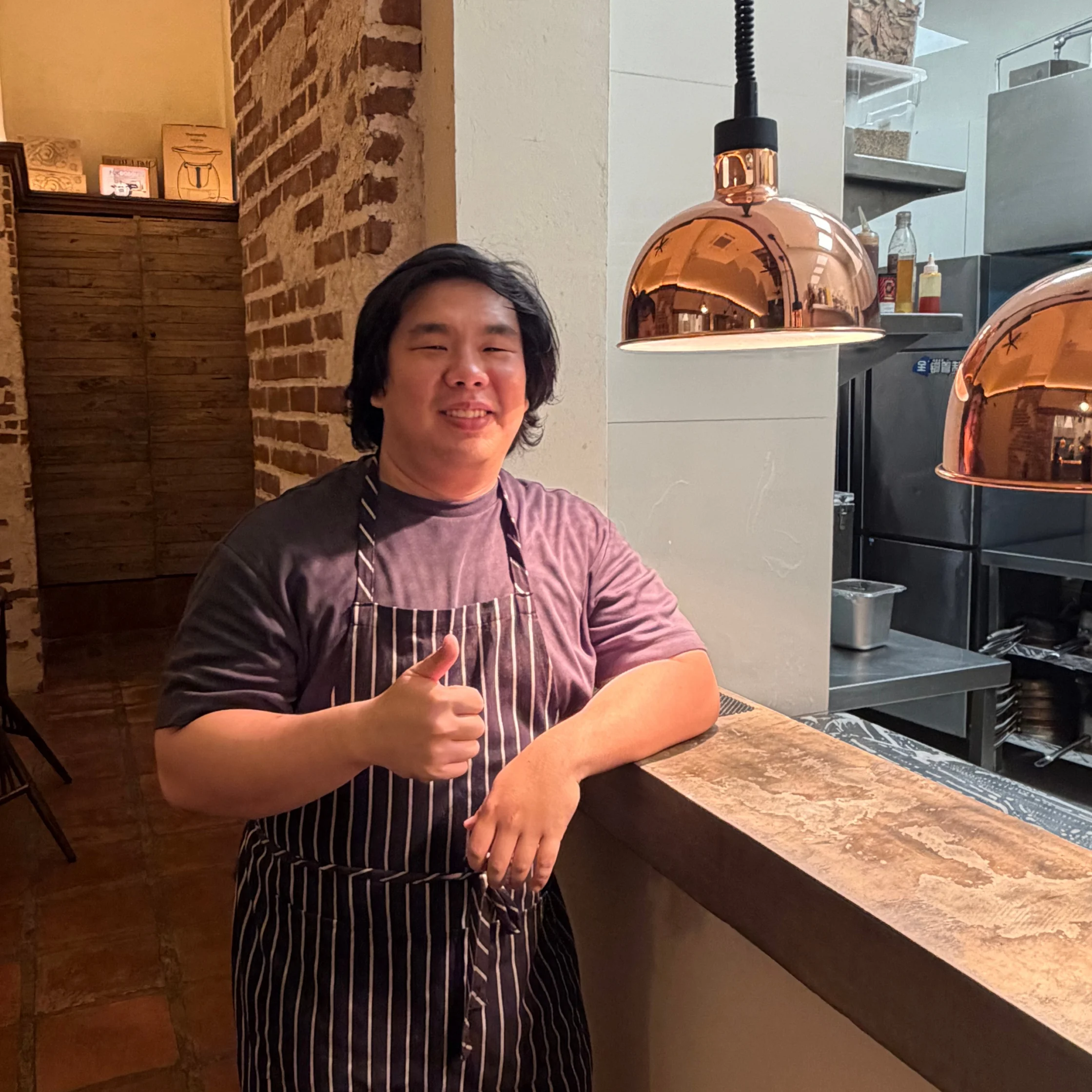 A smiling restaurant worker in a striped apron gives a thumbs-up beside a stone counter, with warm copper pendant lights creating a welcoming atmosphere.