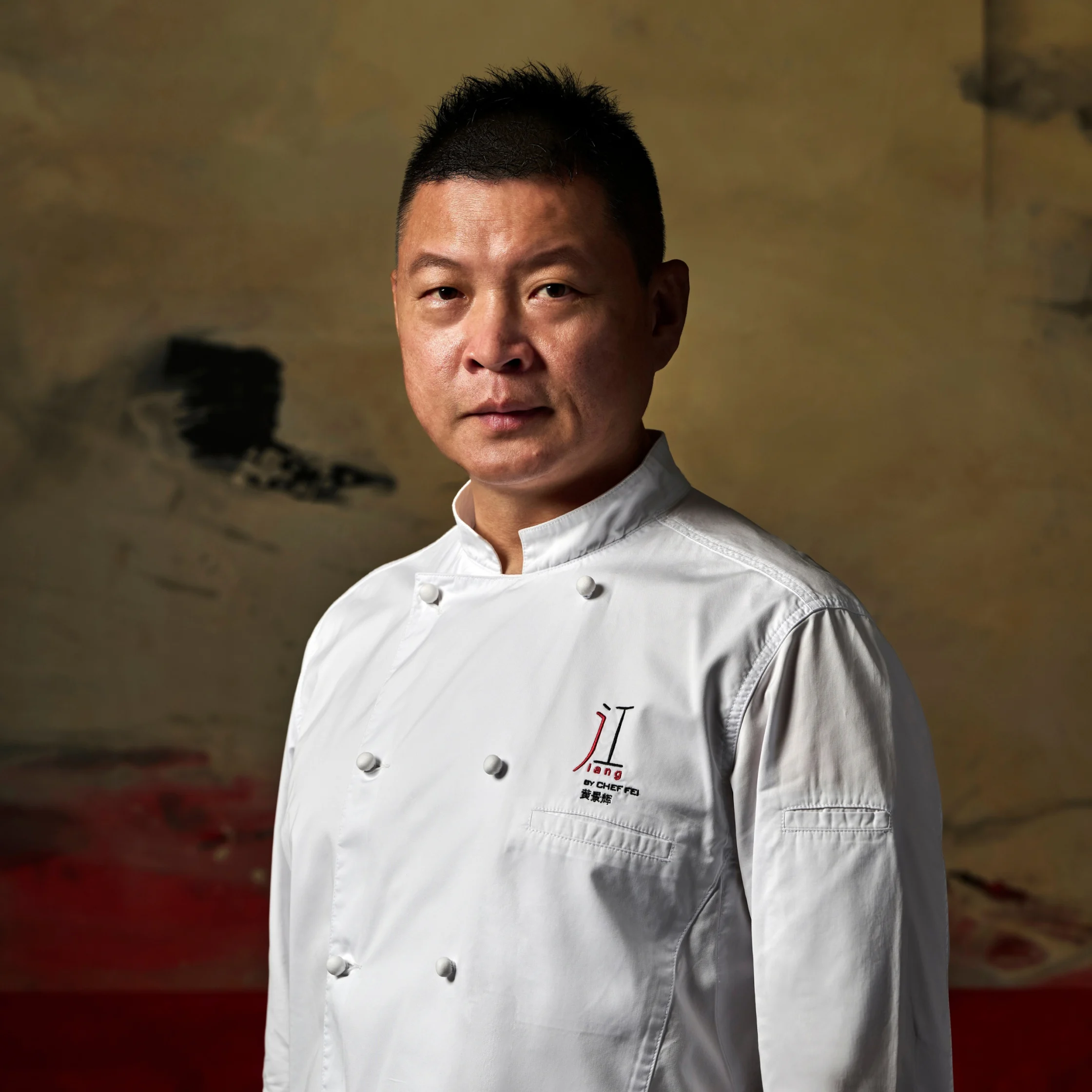 Portrait of a focused male chef in a white double-breasted jacket, standing against a muted, textured wall; the warm lighting feels calm and professional.