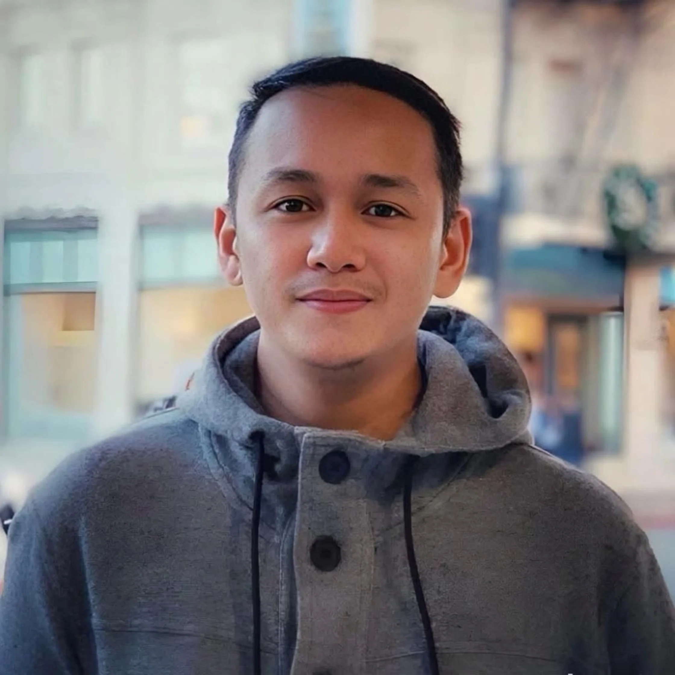 A young man in a gray hooded coat looks directly at the camera with a calm, slight smile, standing against a softly blurred urban background.