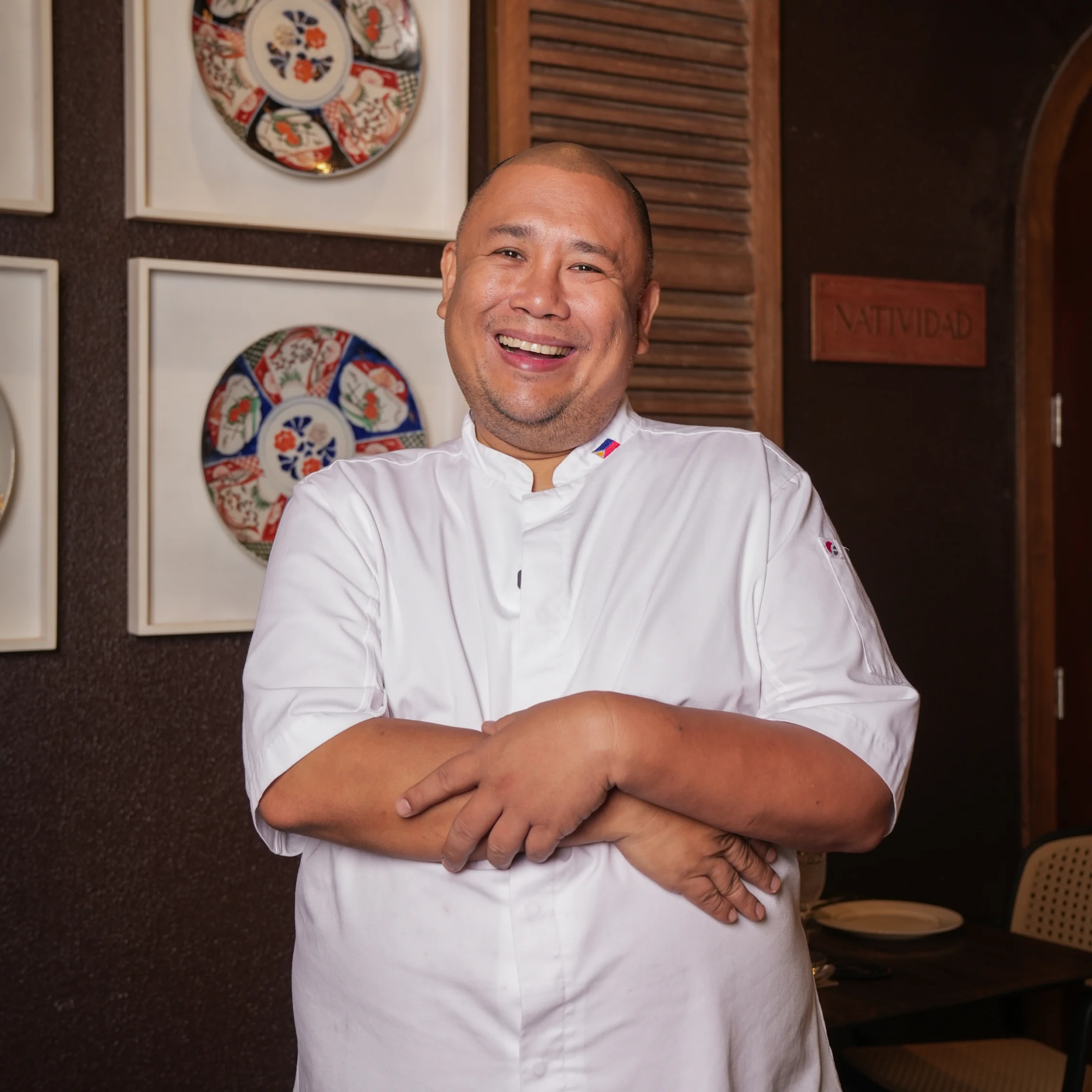 Smiling chef in a white jacket stands with arms crossed in a restaurant, framed by decorative plates and a wooden door; the mood is warm and welcoming.