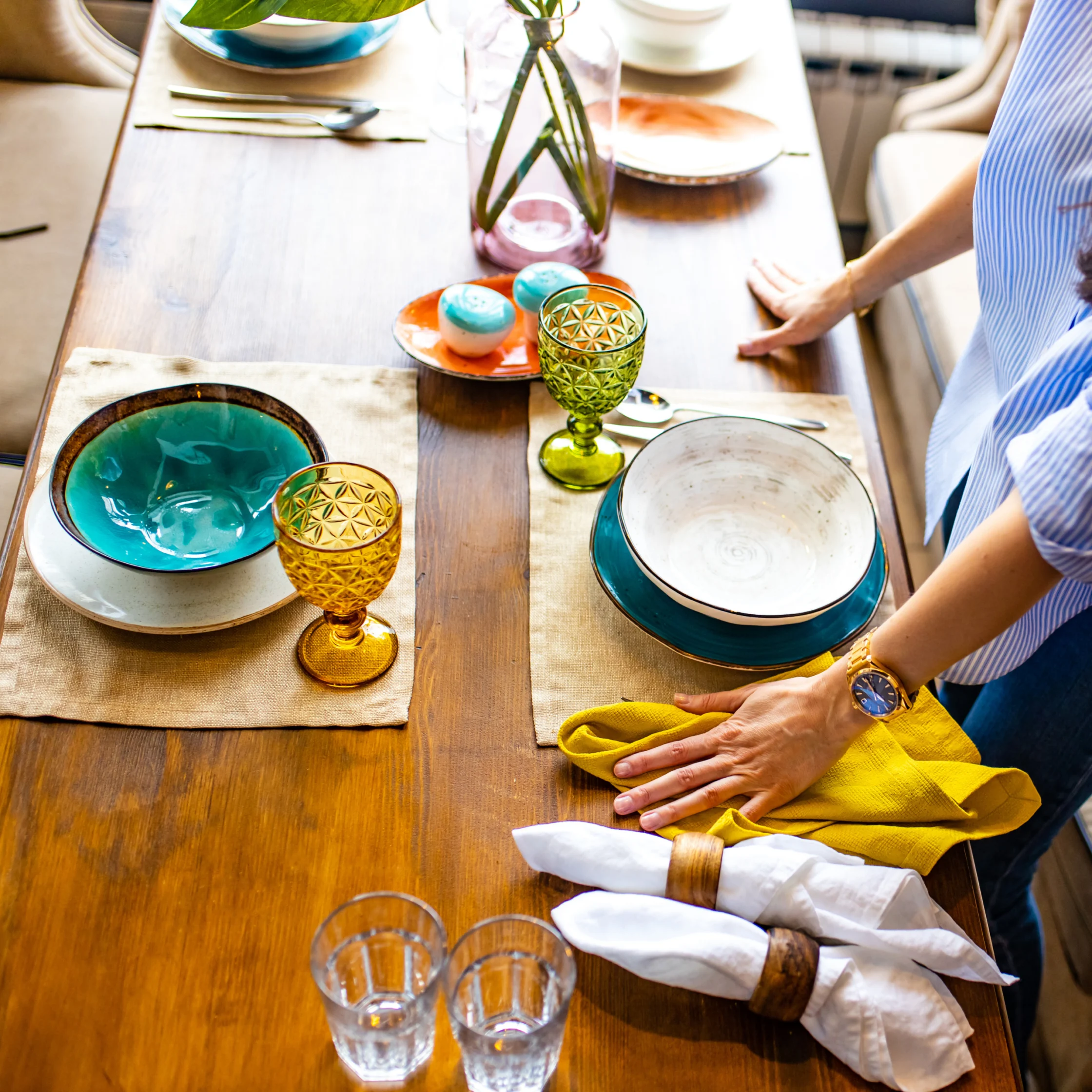 A woman sets a wooden dining table with teal plates, patterned glasses, folded white napkins, and a yellow cloth in a bright, welcoming home.