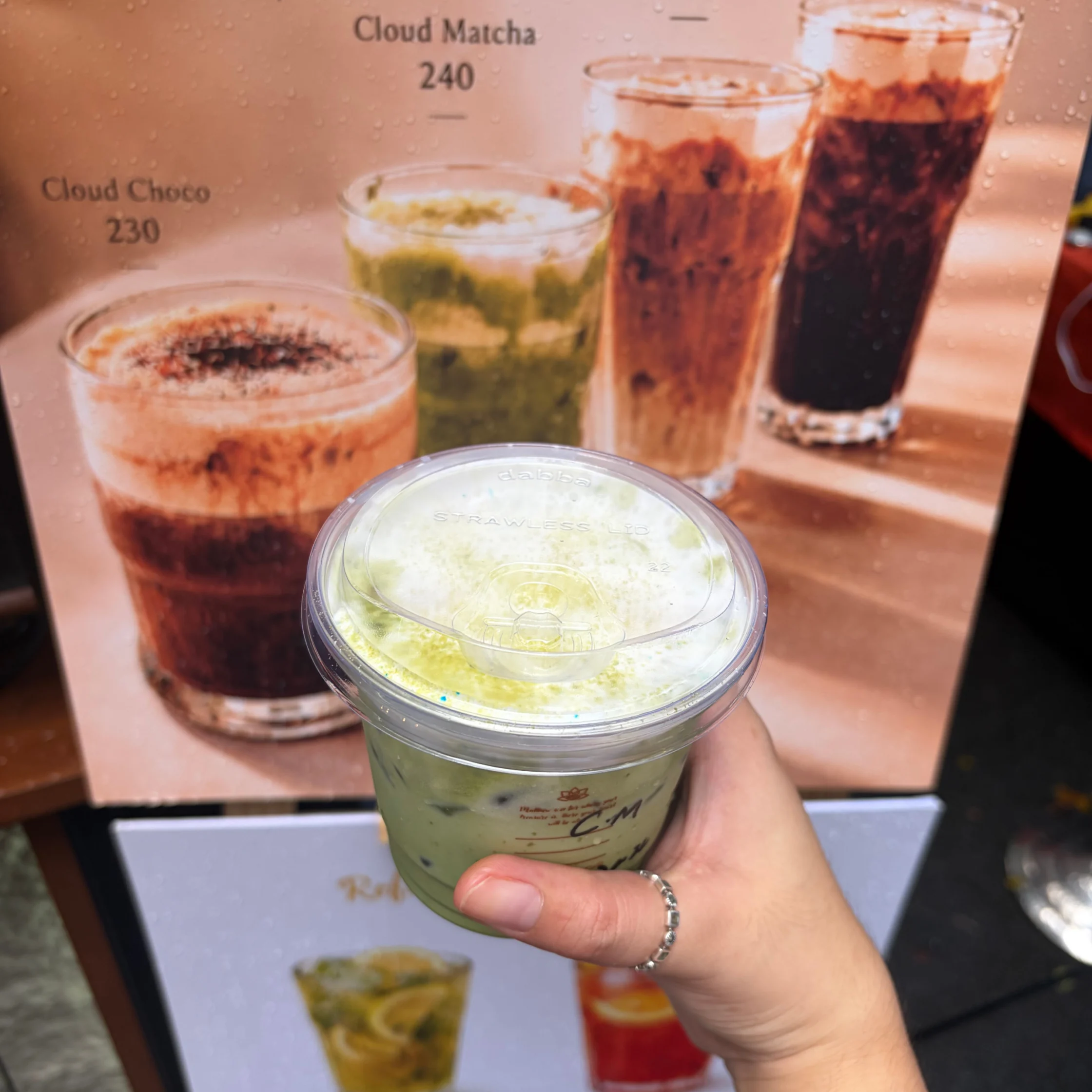 A hand holds a creamy green matcha drink beneath a café menu showing layered Cloud Matcha and Cloud Choco drinks, creating a warm, inviting mood.