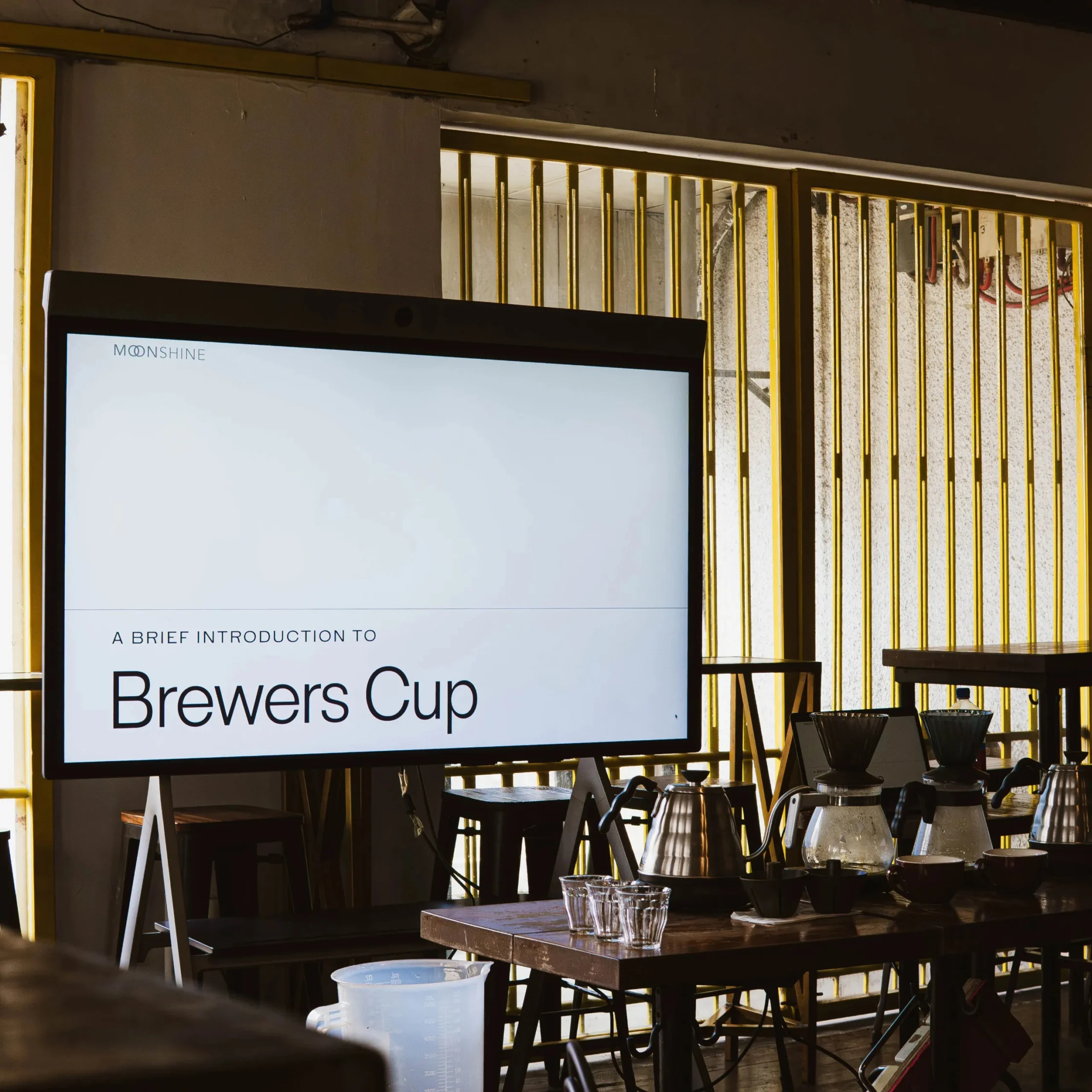 A screen in a warmly lit café displays “A Brief Introduction to Brewers Cup,” with coffee brewers and glassware on a nearby table; the mood is calm and educational.