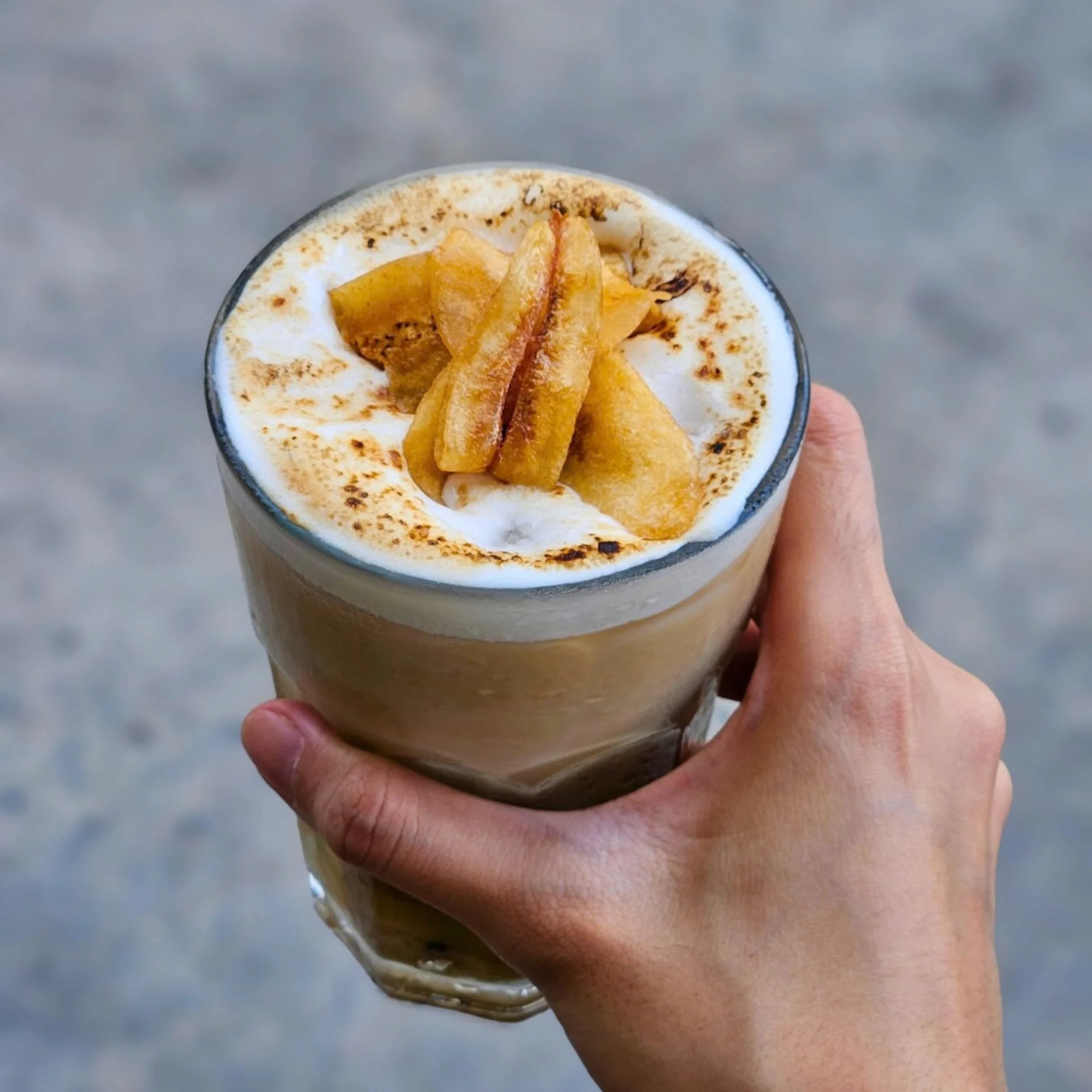 A hand holds a layered latte topped with thick foam, caramelized banana slices, and cinnamon, offering a cozy, indulgent café treat against a cool gray background.