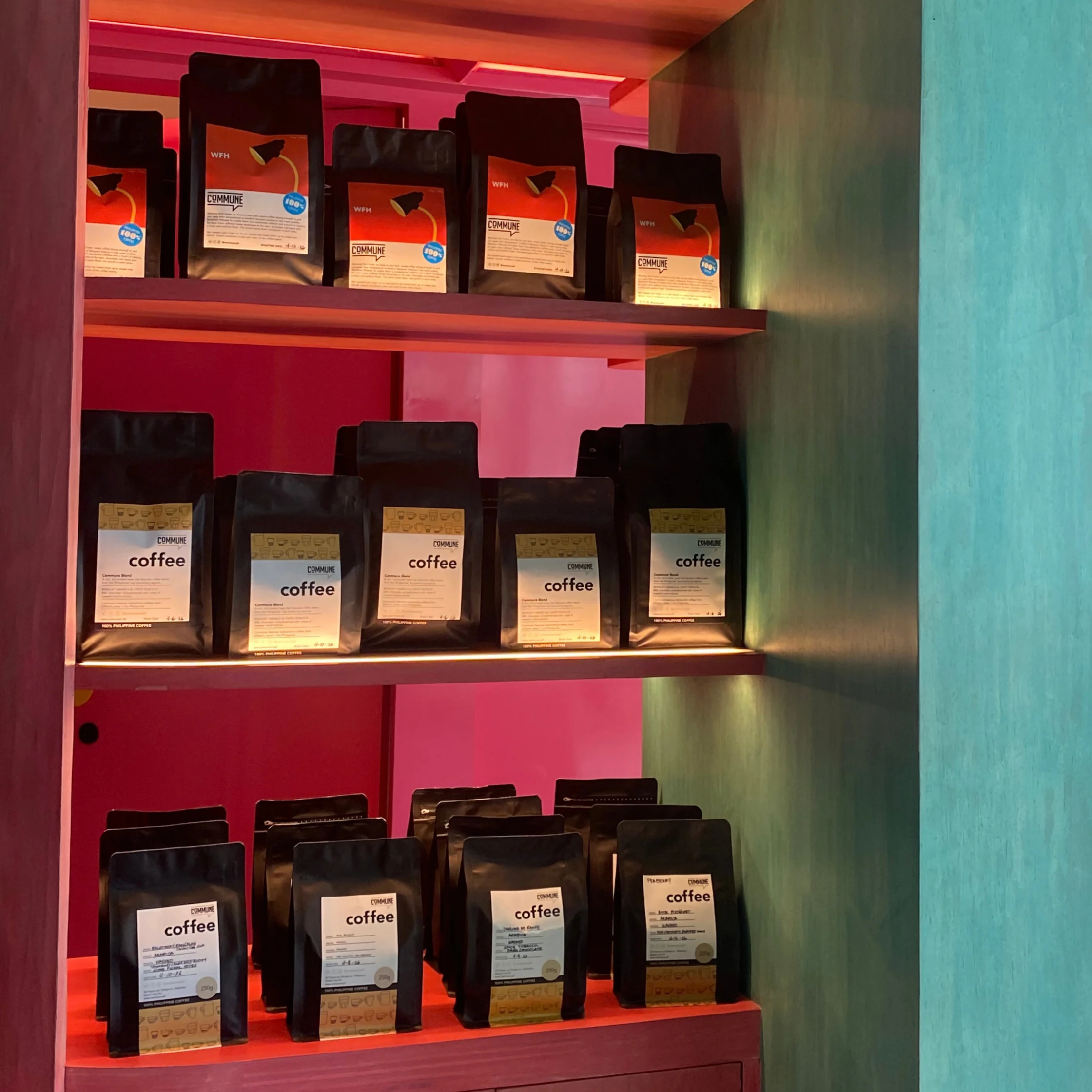 Rows of packaged coffee sit on red-lit wooden shelves in a teal cabinet, creating a vivid, cozy specialty-shop display with a warm, inviting feel.