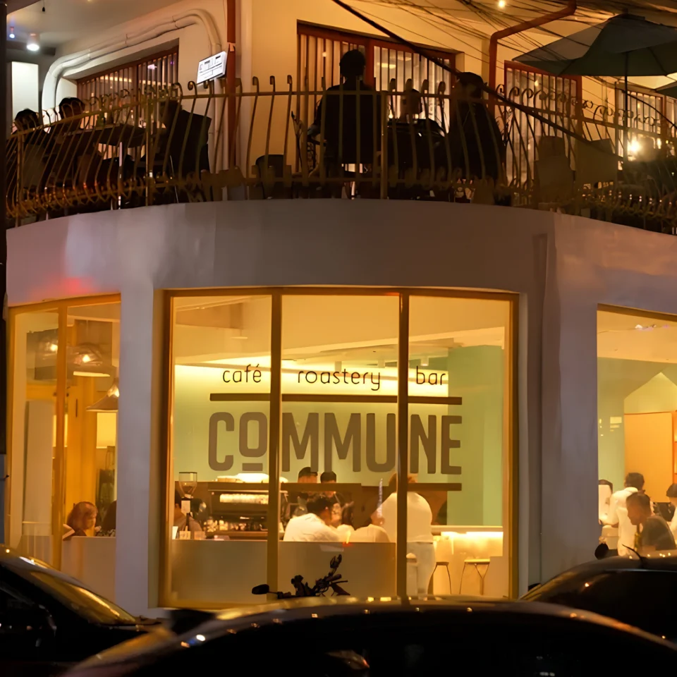 Nighttime street view of Commune café, roastery, and bar, glowing warmly through large windows as patrons gather inside and on the balcony above.