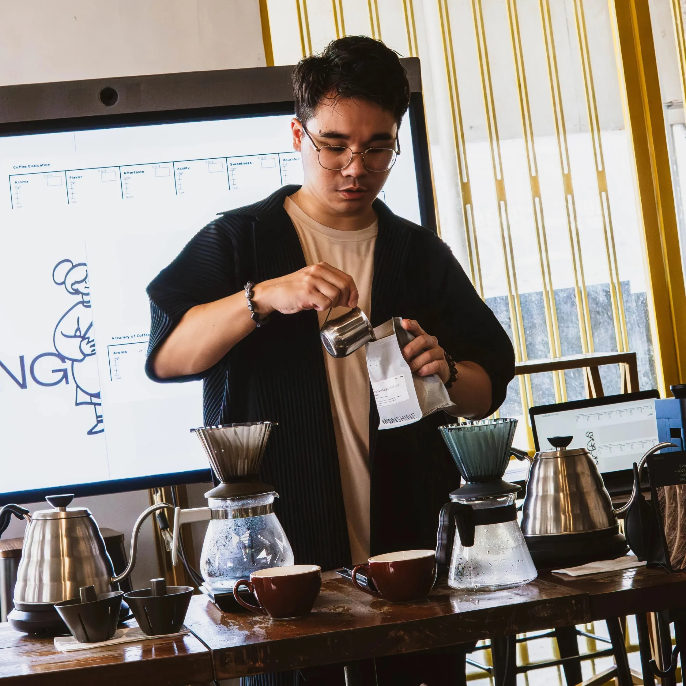 A barista pours freshly ground coffee from a metal scoop into a filter at a bright café brewing station, focused amid kettles and drippers.