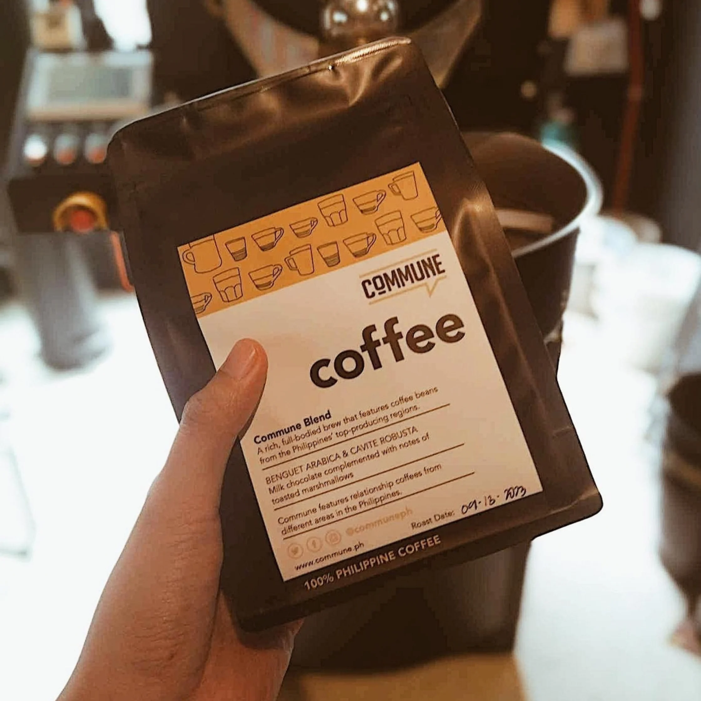 A hand holds a dark bag of Commune coffee labeled “100% Philippine Coffee” in a warmly lit café, with brewing equipment blurred behind it.