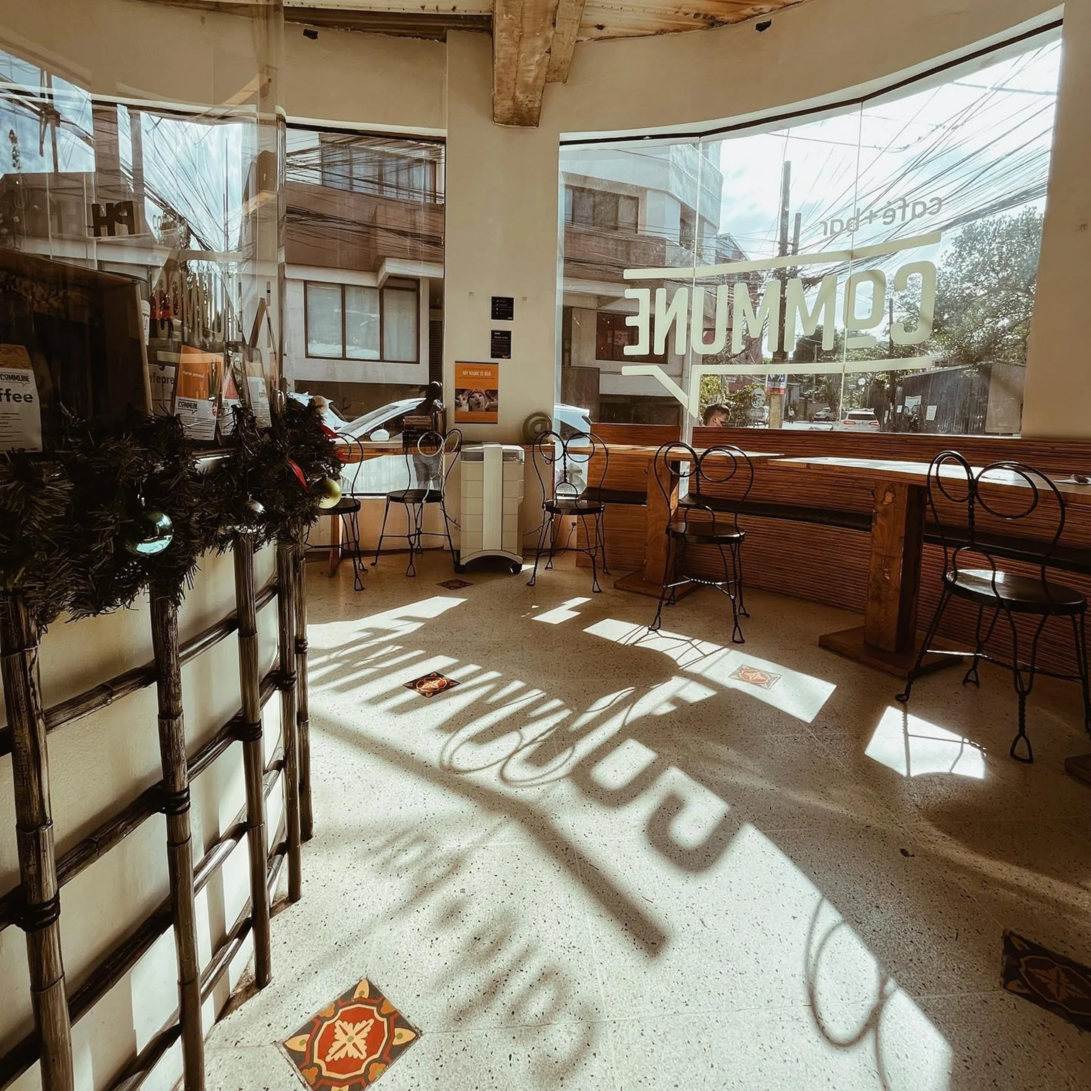 Sunlit café interior with metal chairs, a long wooden counter, and large windows; reversed “COMING” lettering casts shadows across the floor, creating a quiet, inviting mood.