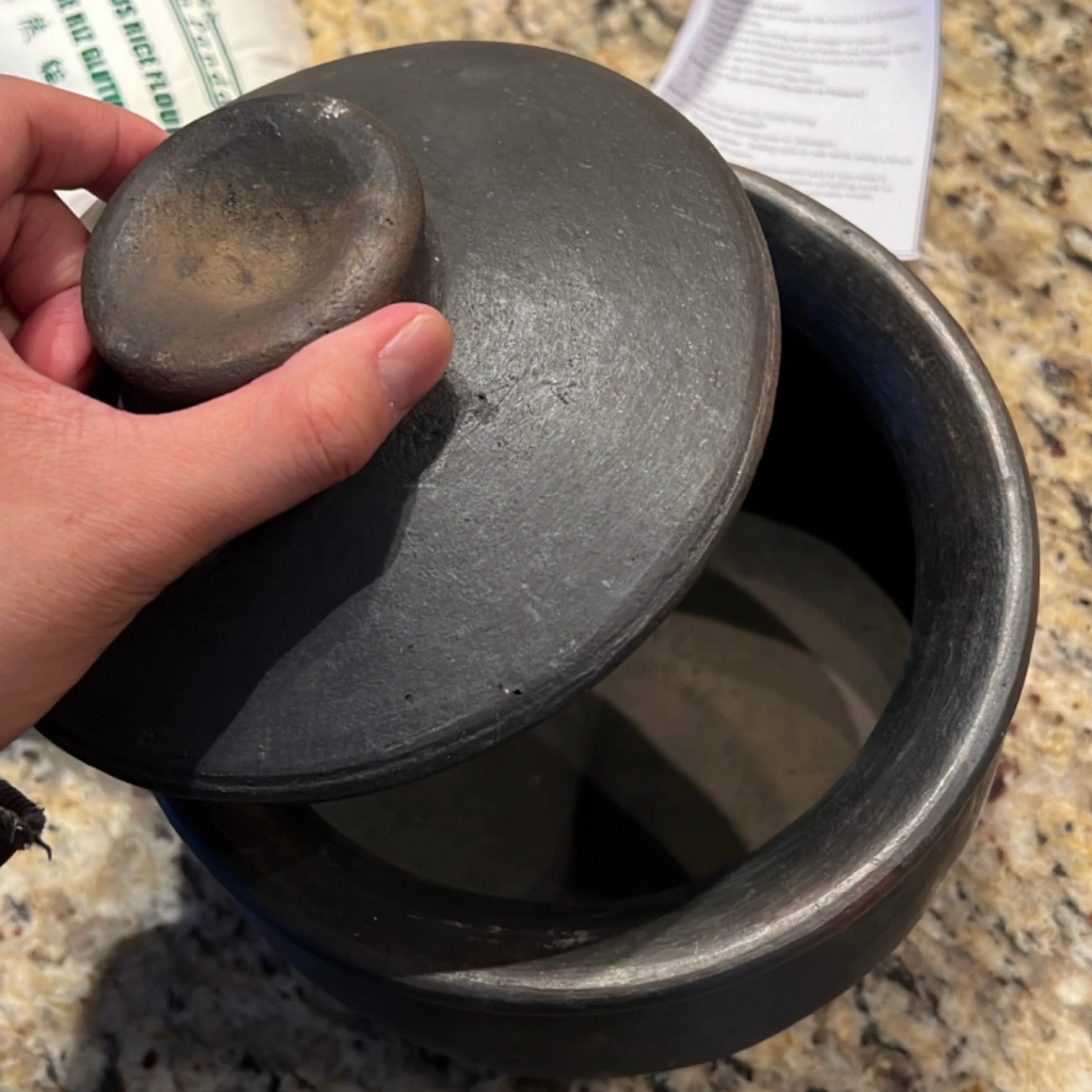 A hand lifts the heavy lid of a dark stone cooking pot, revealing its empty interior on a speckled counter; the scene feels rustic and practical.
