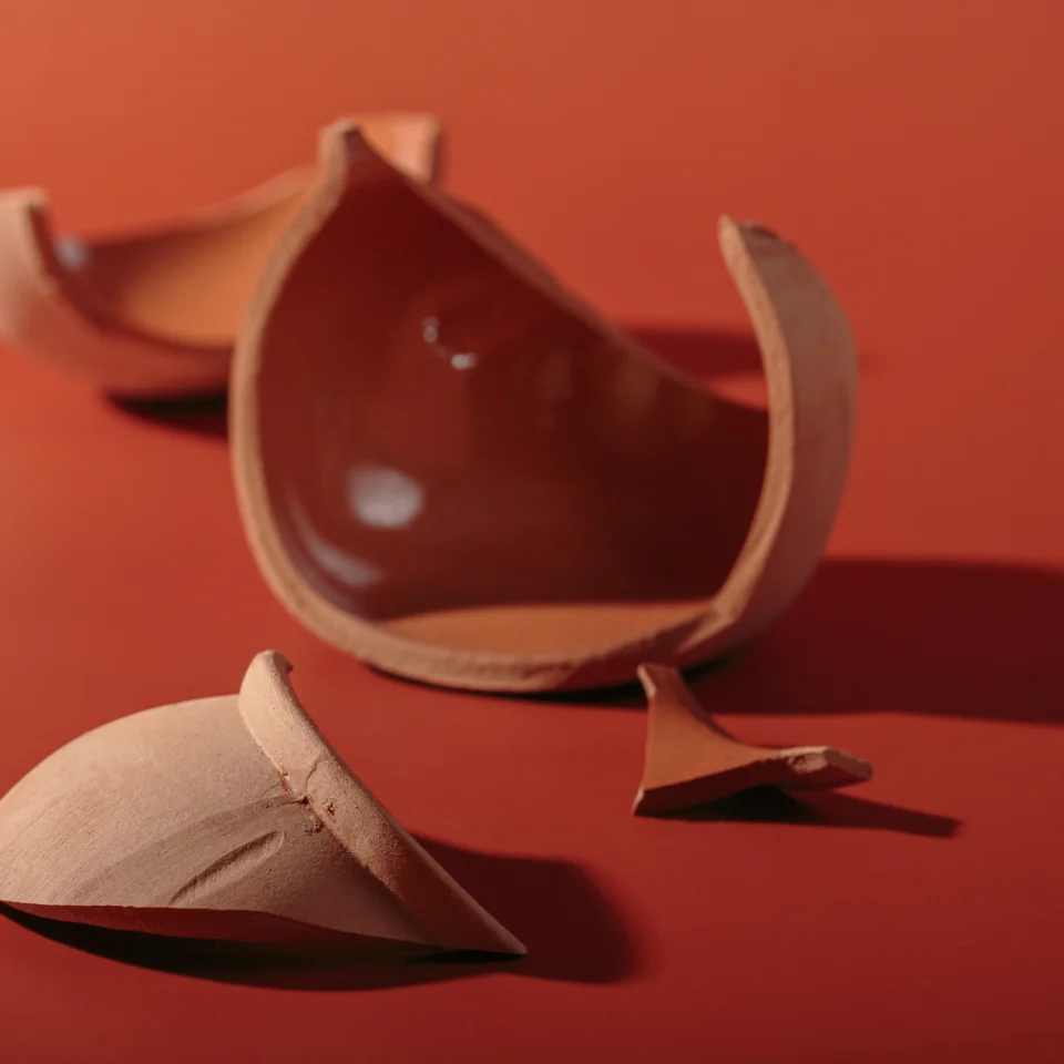 Broken terracotta pottery pieces—a glossy curved bowl fragment and shards—rest on a red surface in warm light, creating a quiet, sculptural mood.