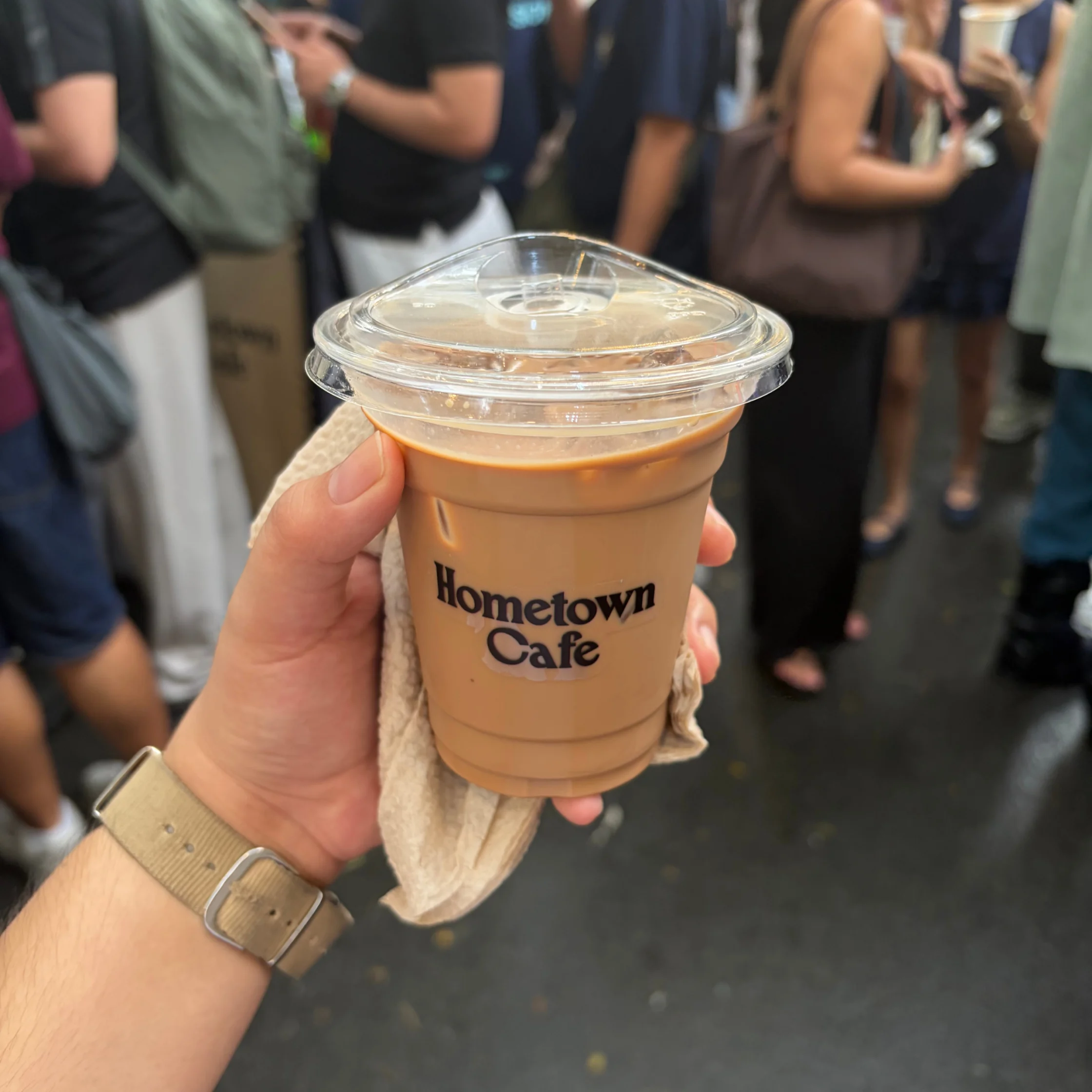 A hand holds a tan iced coffee in a clear-lidded cup labeled “Hometown Cafe,” amid a busy crowd; the casual café scene feels lively and welcoming.