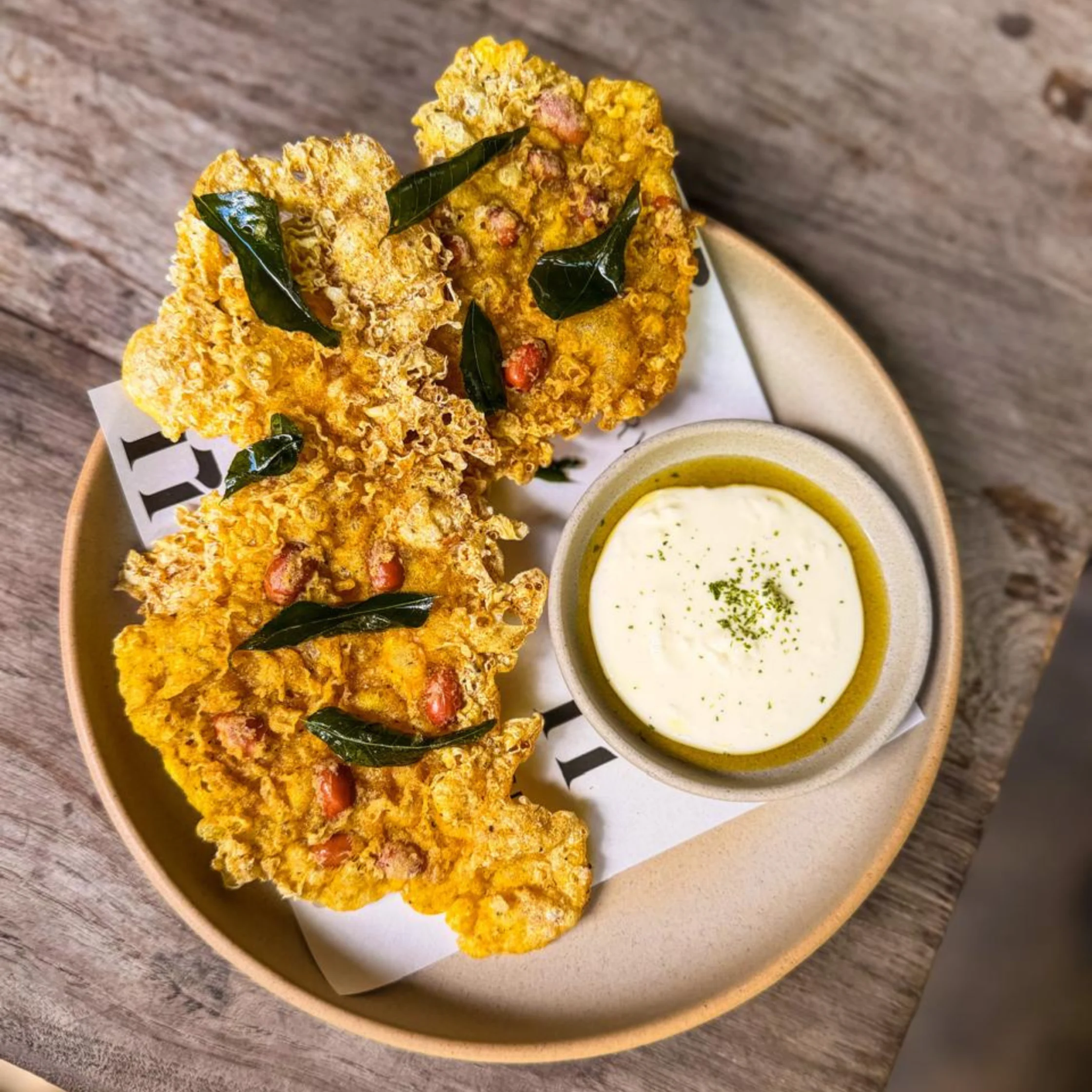 Golden, crispy fritters topped with curry leaves and red peanuts sit on a plate beside creamy dipping sauce, evoking a warm, savory snack.