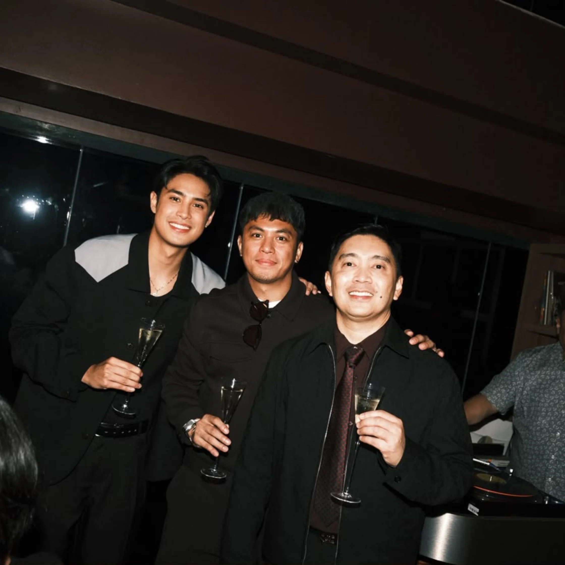 Three men in dark suits pose closely at a dimly lit indoor gathering, smiling and holding drinks; their warm, celebratory mood stands out against the dark background.