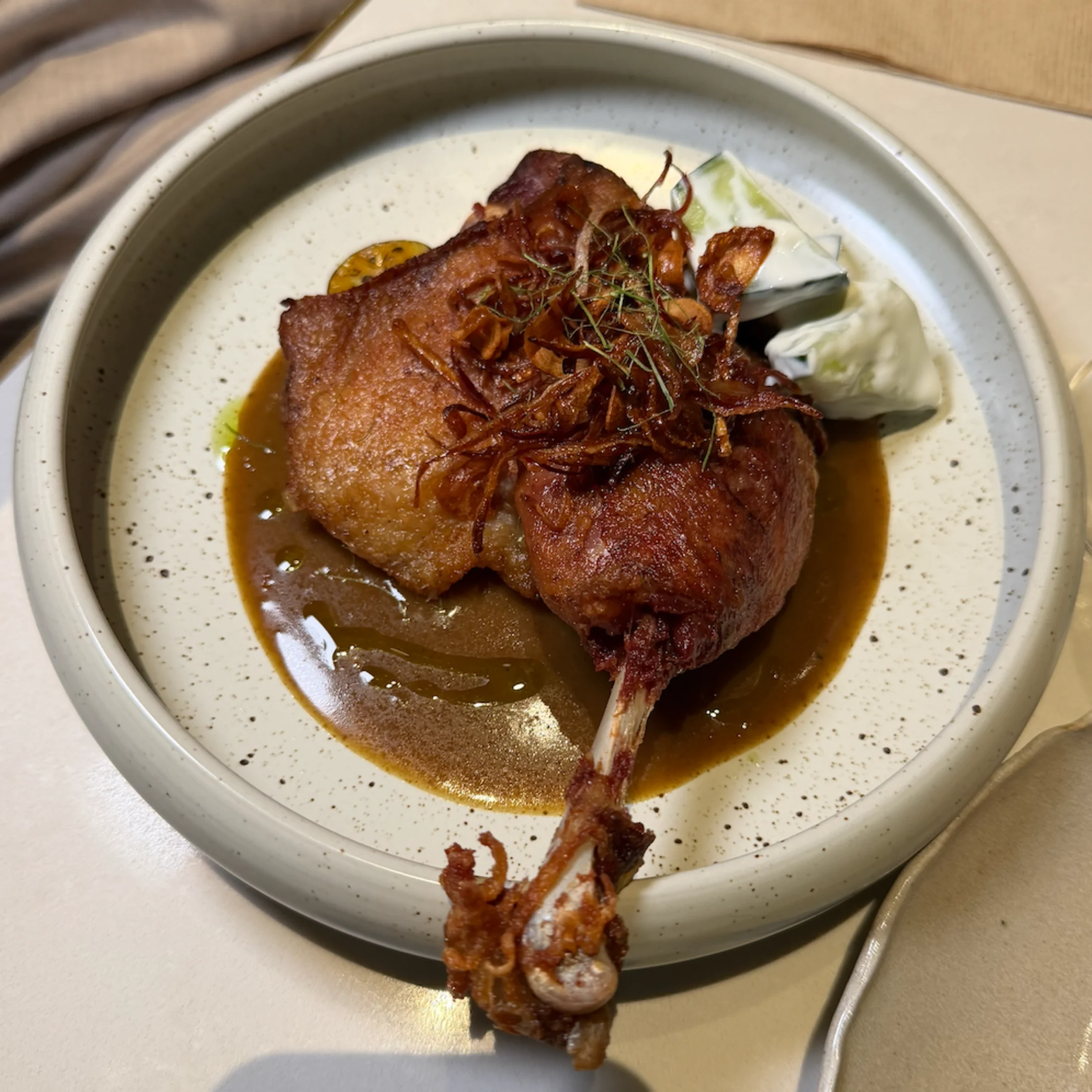 Golden-brown roasted duck leg with crisp skin sits in glossy brown gravy, garnished with herbs and served with pale cabbage on a ceramic plate; warm, rustic dining.