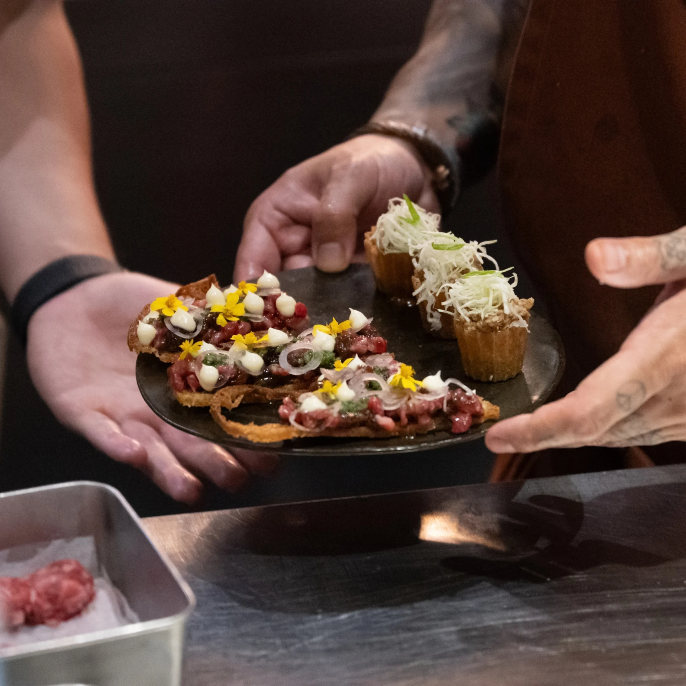 Hands serve a black platter of gourmet appetizers—tartines topped with meat, creamy bites and edible flowers, plus shredded-topped cups—creating an elegant dining mood.