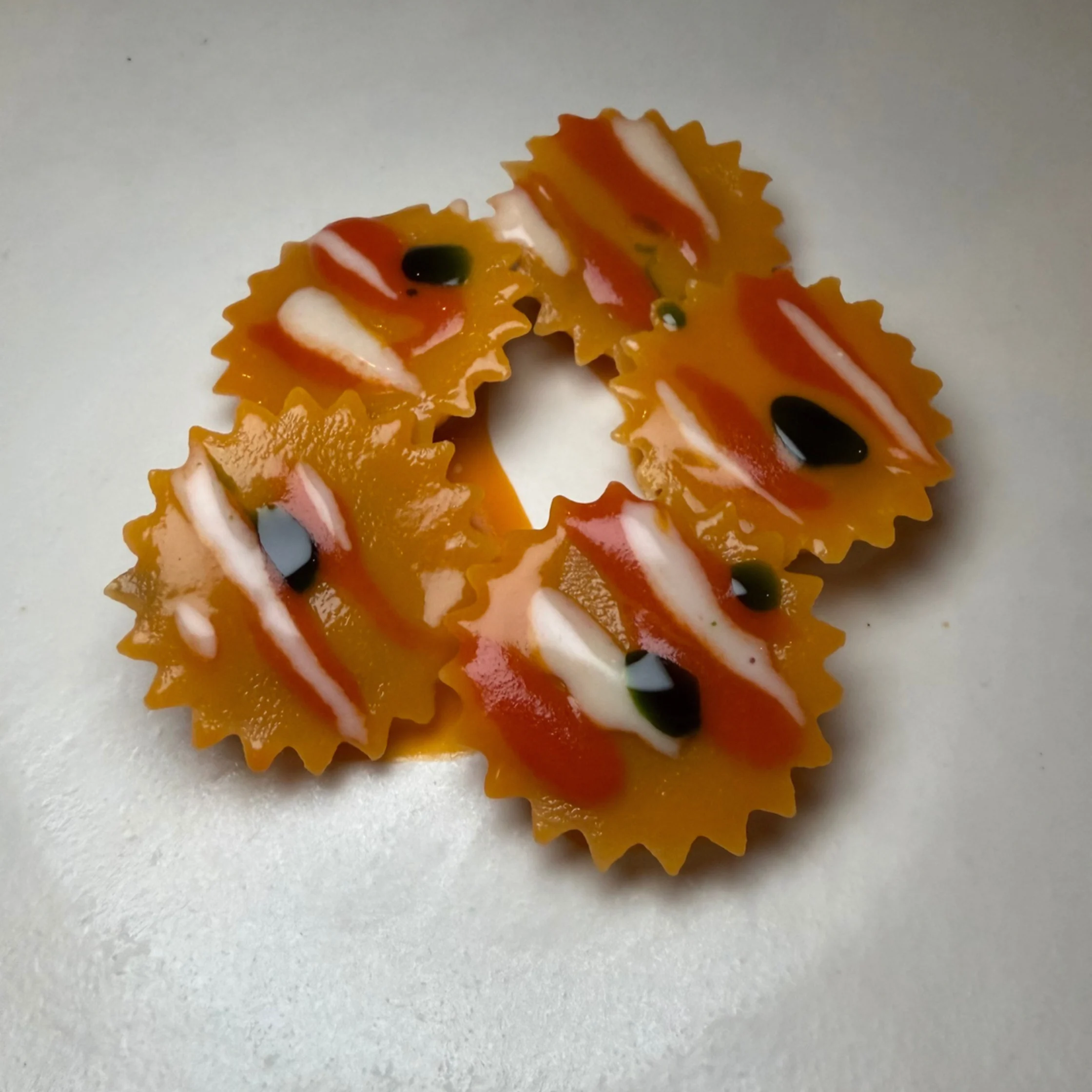 Four colorful, flower-shaped pasta pieces with orange-red sauces, cream streaks, and dark accents sit on a pale surface, creating a playful, appetizing look.