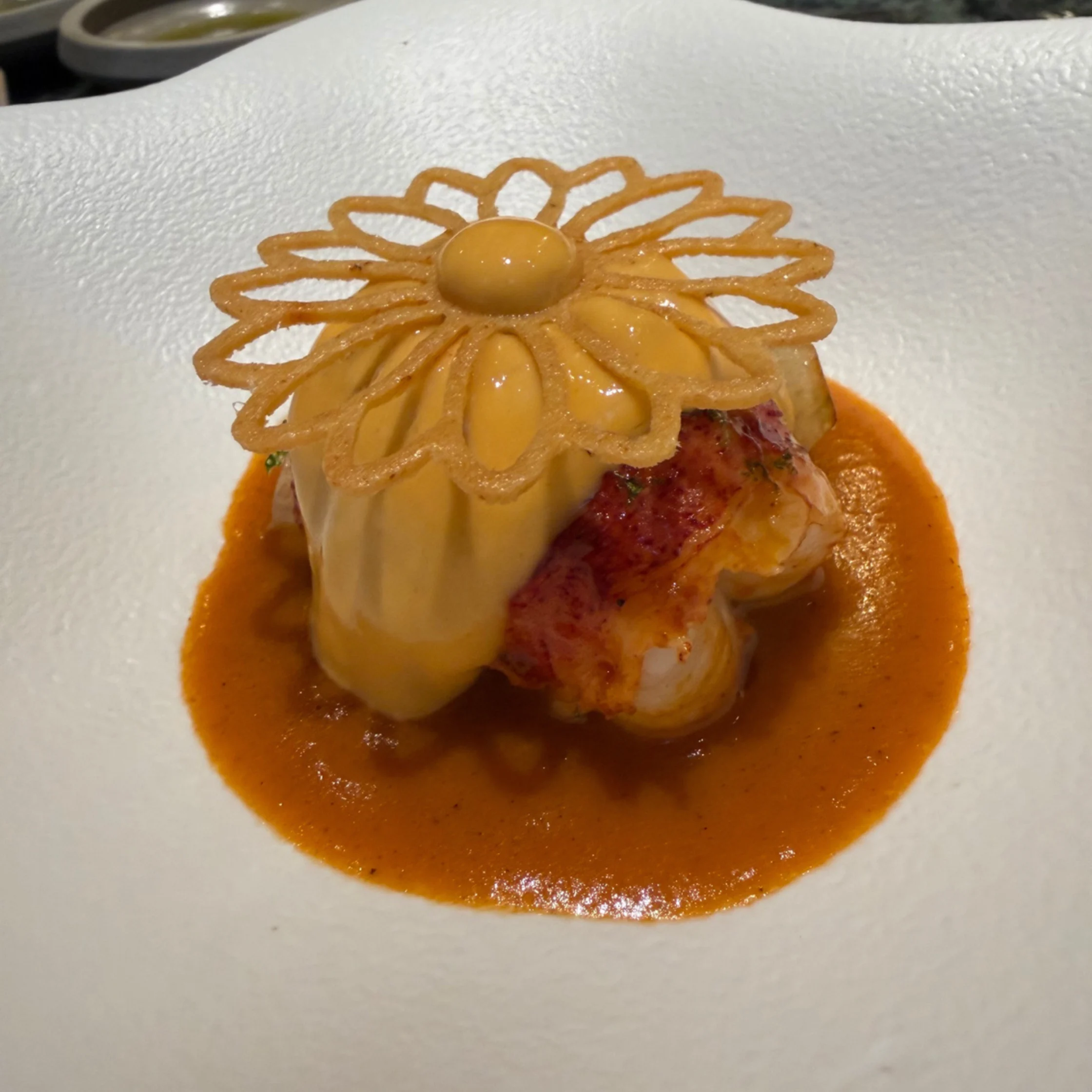 A refined seafood dish on a white plate: lobster sits in a pool of orange sauce beneath a flower-shaped crisp garnish, creating an elegant restaurant presentation.