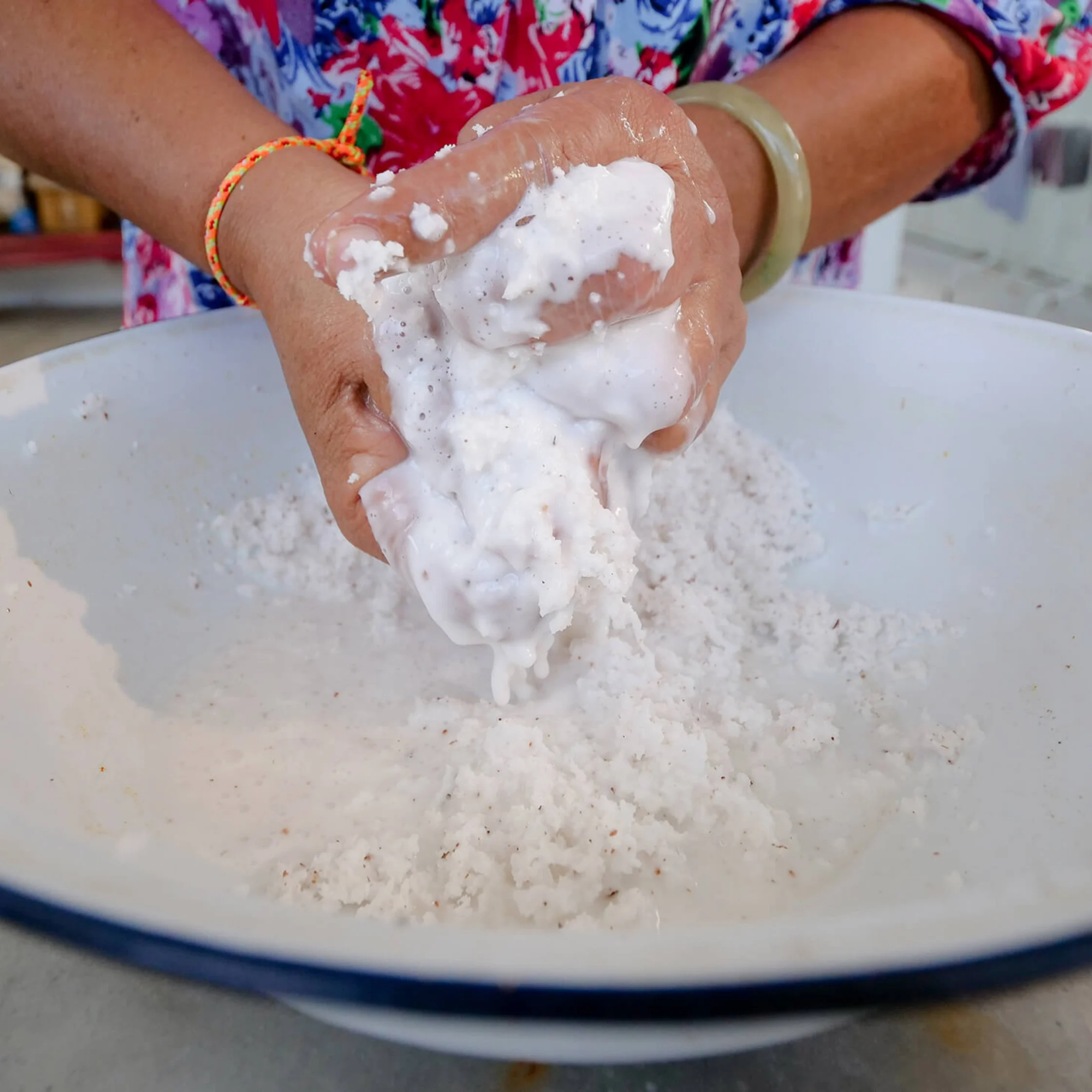 Hands squeeze a wet white coconut mixture over a large basin, with liquid and shreds dripping down; colorful clothing adds a lively, hands-on feel.