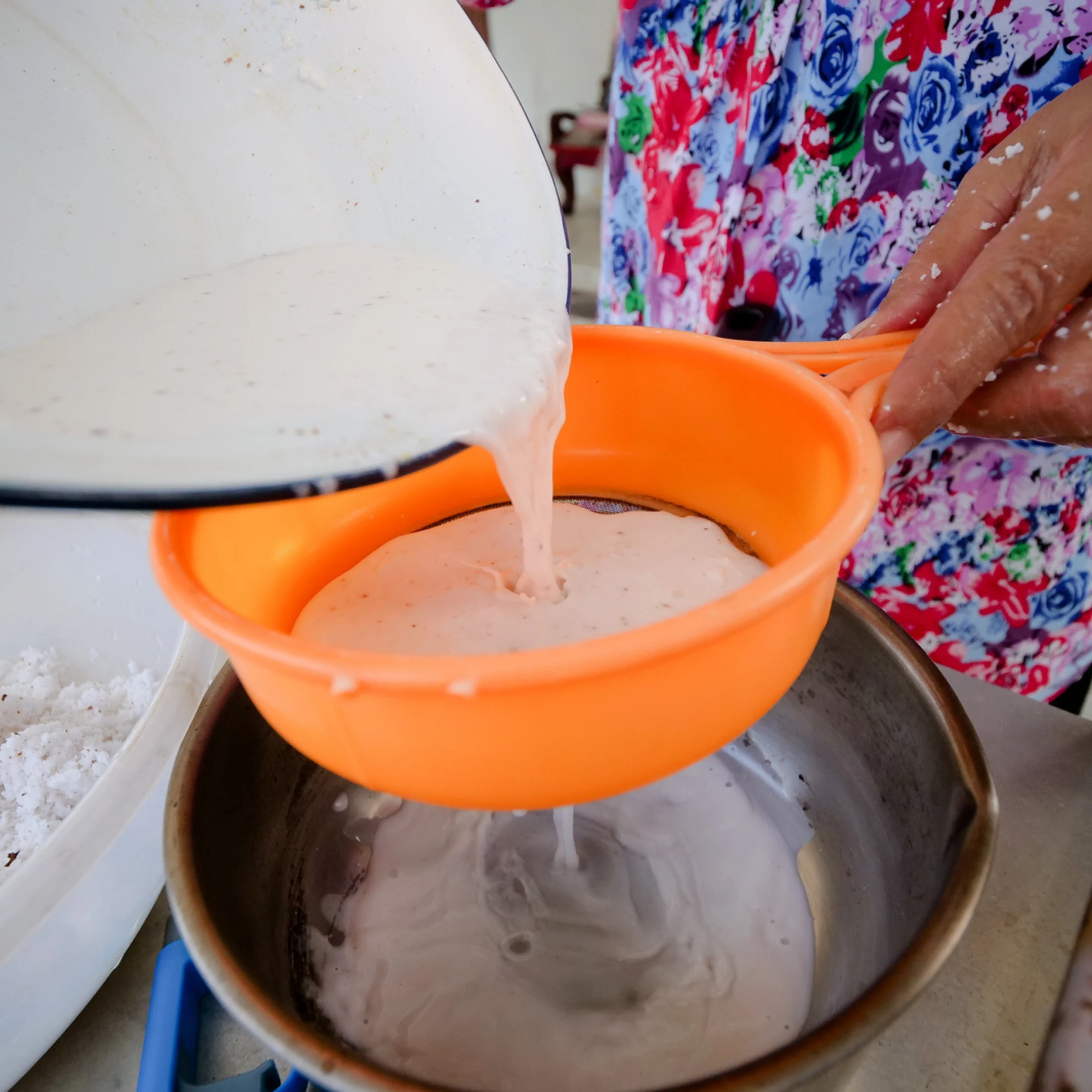 A cook pours creamy batter through an orange strainer into a metal bowl, with a floral apron visible in this focused home-kitchen scene.