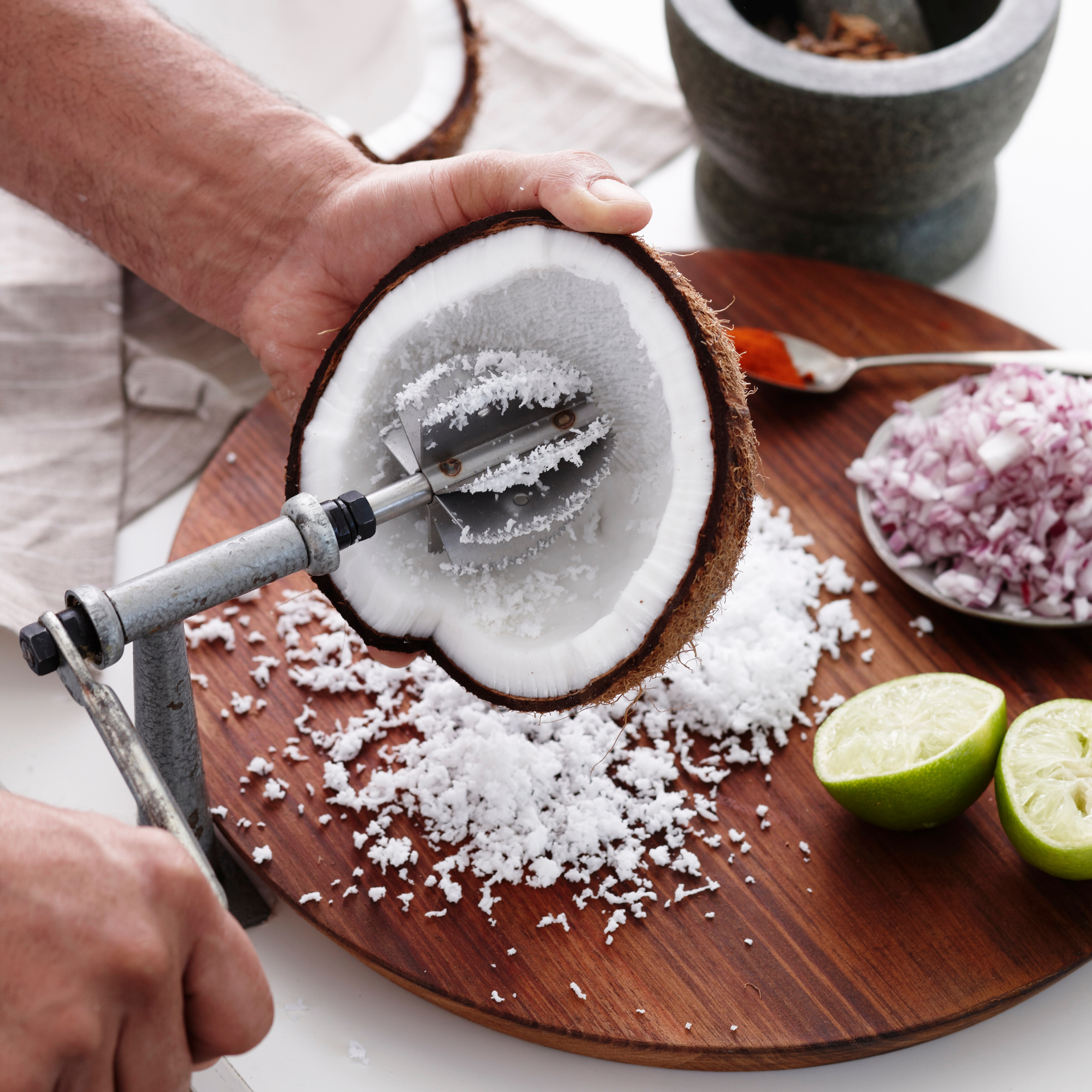 Hands grate fresh coconut over a wooden board, with shredded coconut, chopped onion, lime, and mortar nearby; the scene feels rustic and hands-on.