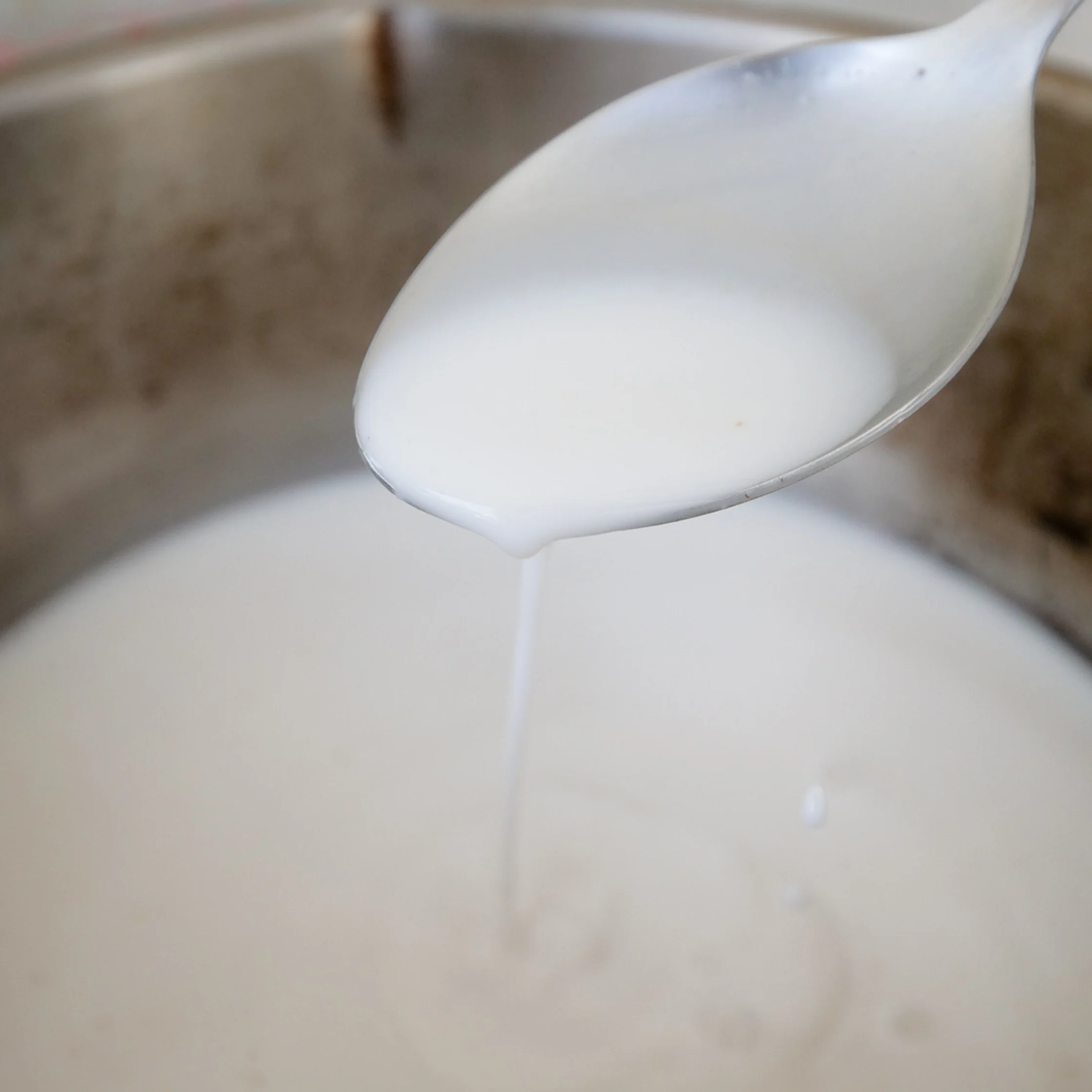 A white spoon lifts creamy batter above a metal mixing bowl as a thin stream drips back, showing its smooth consistency in a calm, close-up kitchen scene.