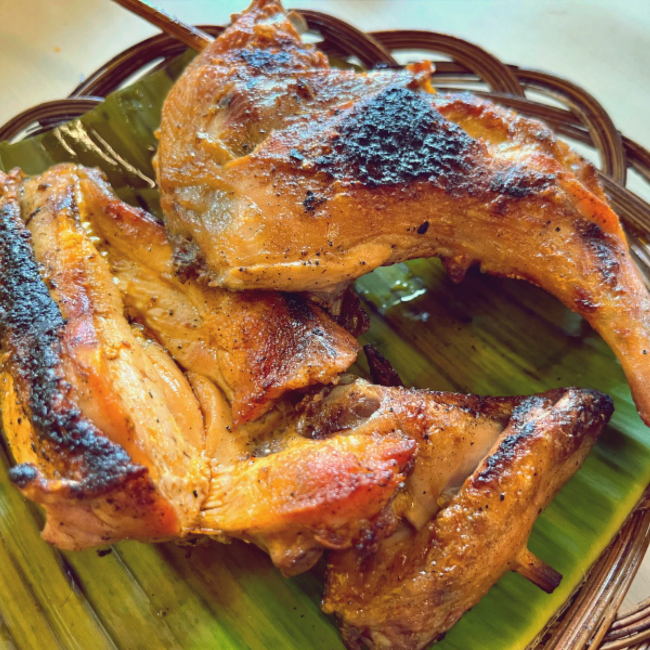 Golden, char-grilled chicken pieces with dark, smoky grill marks rest on glossy banana leaves in a woven basket, evoking a hearty, appetizing meal.