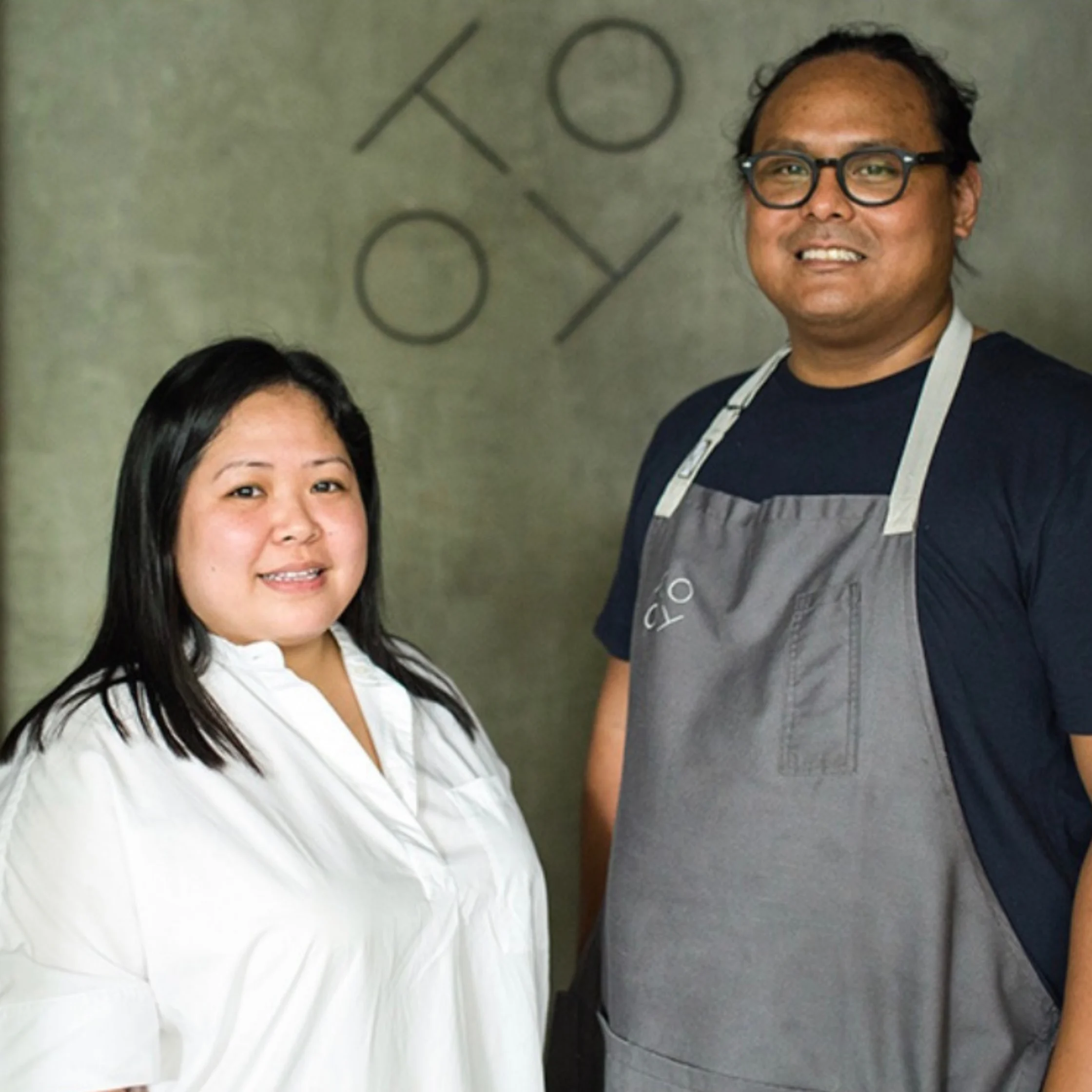 Two smiling restaurant staff members pose against a concrete wall marked “KOYO”: a woman in a white shirt and a man wearing glasses and a gray apron.