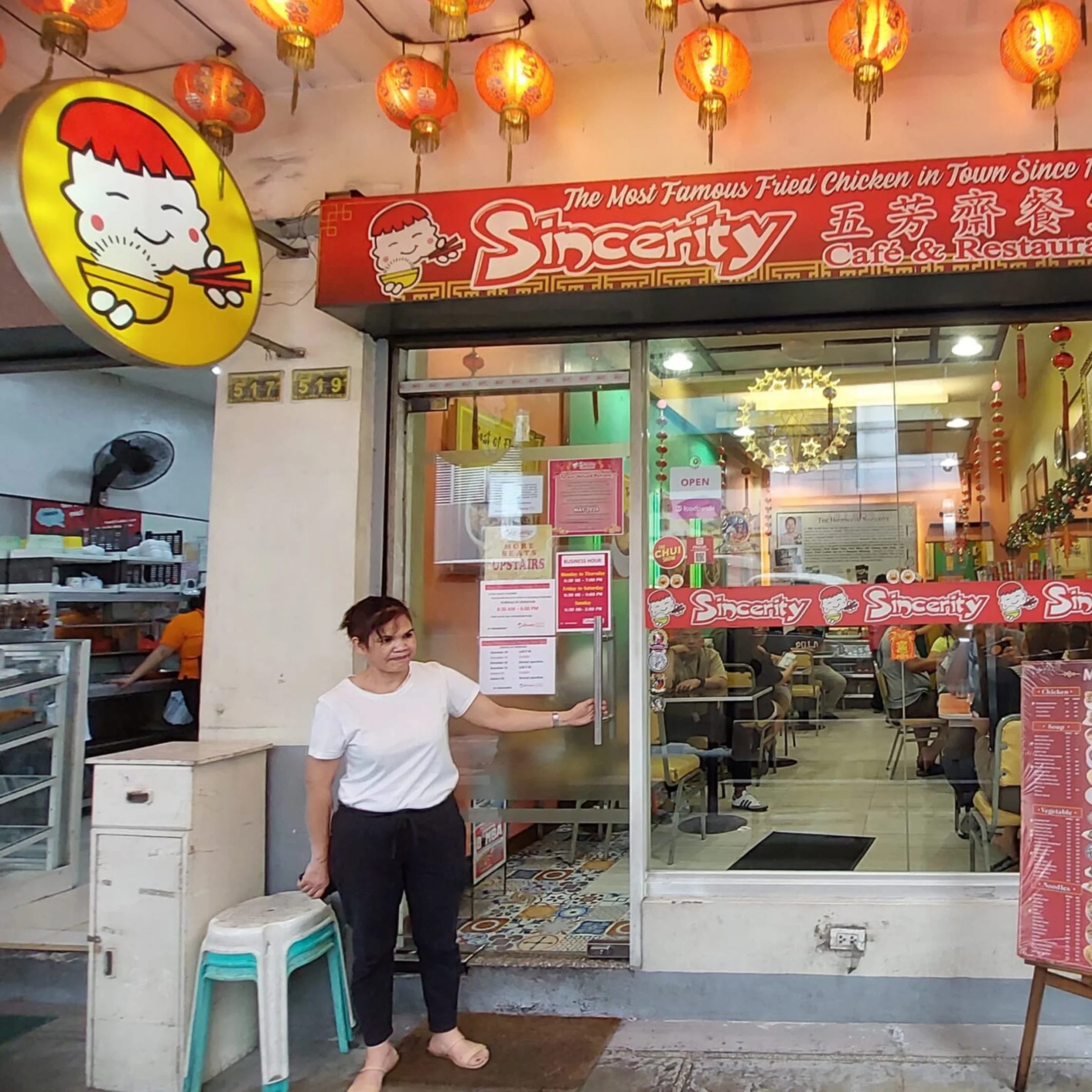 A woman holds open the glass door of Sincerity Chinese fried chicken restaurant beneath glowing red lanterns, welcoming customers into a bright, casual dining room.
