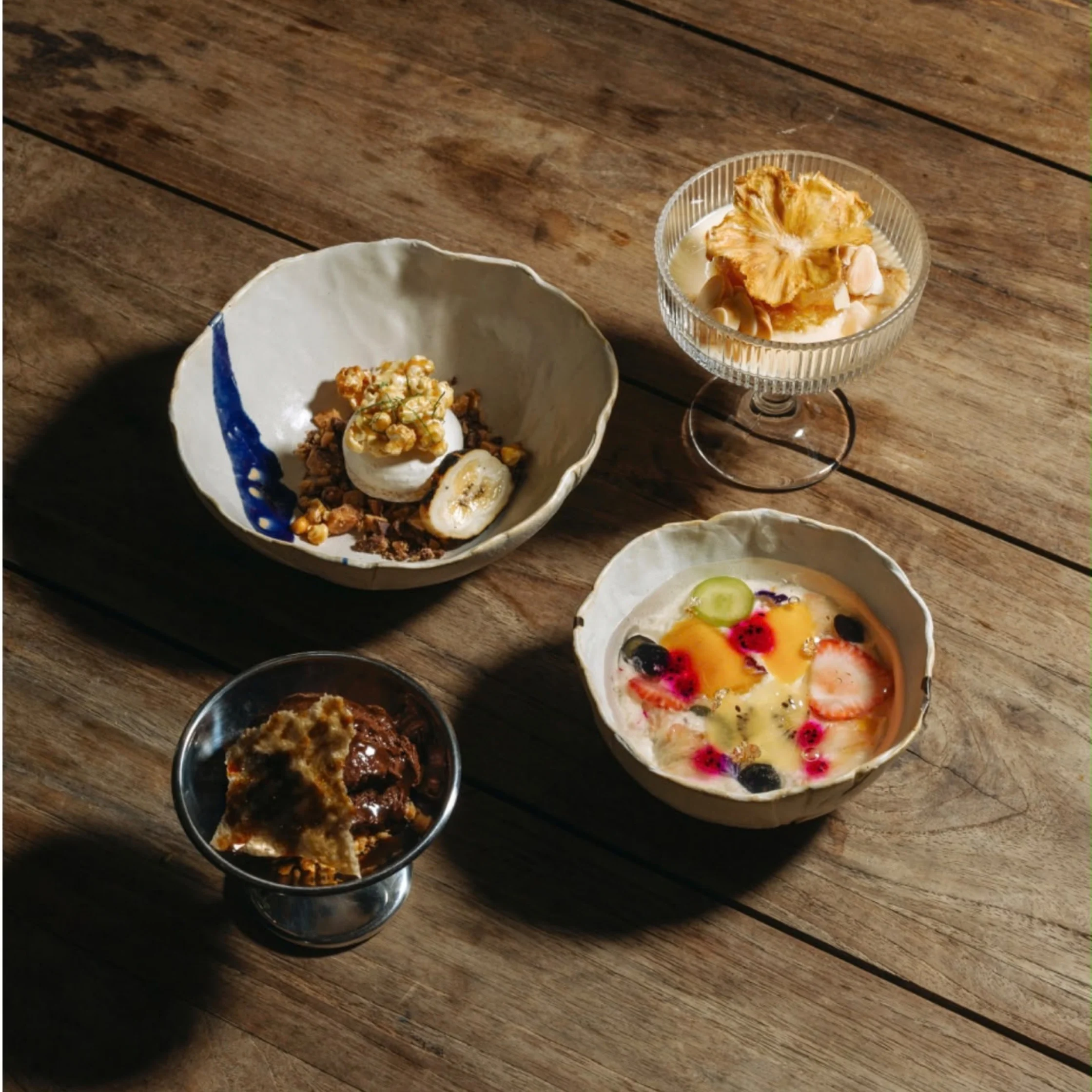 Four artfully plated desserts—colorful fruit in a shallow bowl, layered sweets in a glass coupe, and two rustic dishes—sit on a sunlit wooden table, evoking a warm, inviting mood.