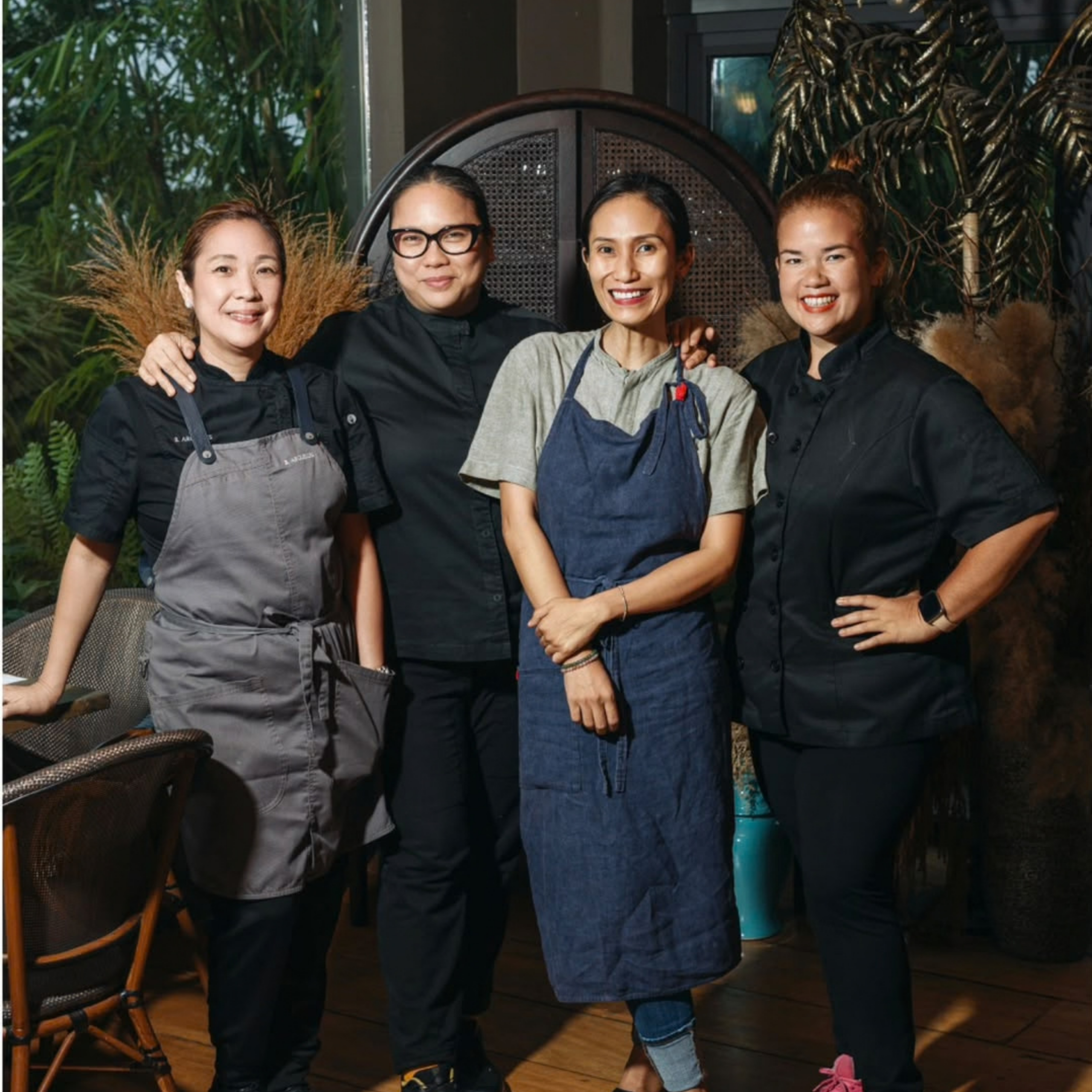 Four smiling women chefs stand together in a warmly lit restaurant, wearing dark work clothes and gray or blue aprons, conveying camaraderie and pride.