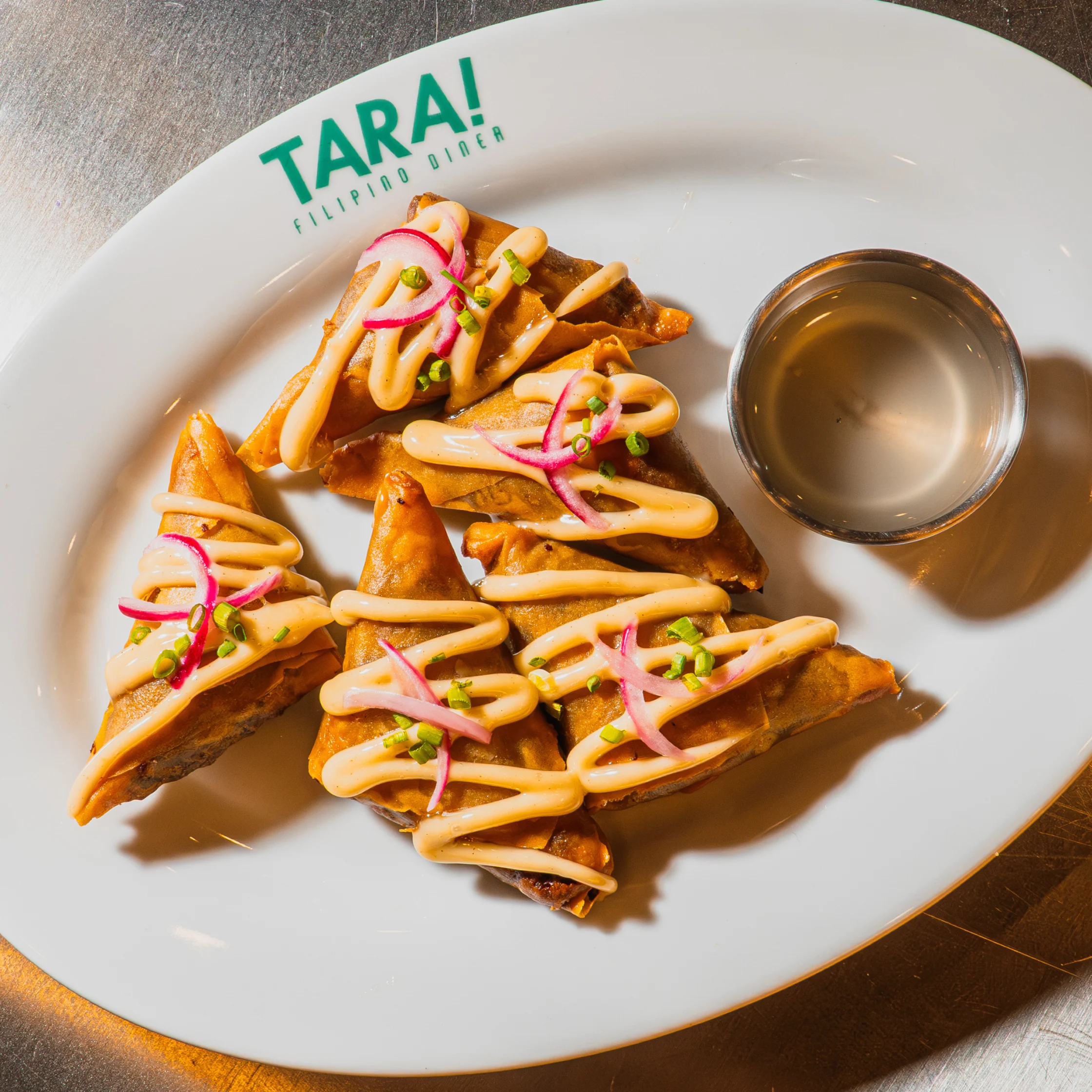 Golden fried samosas drizzled with creamy sauce and topped with pickled onions and herbs sit beside a dipping cup on a white TARA! plate; appetizing, bright presentation.