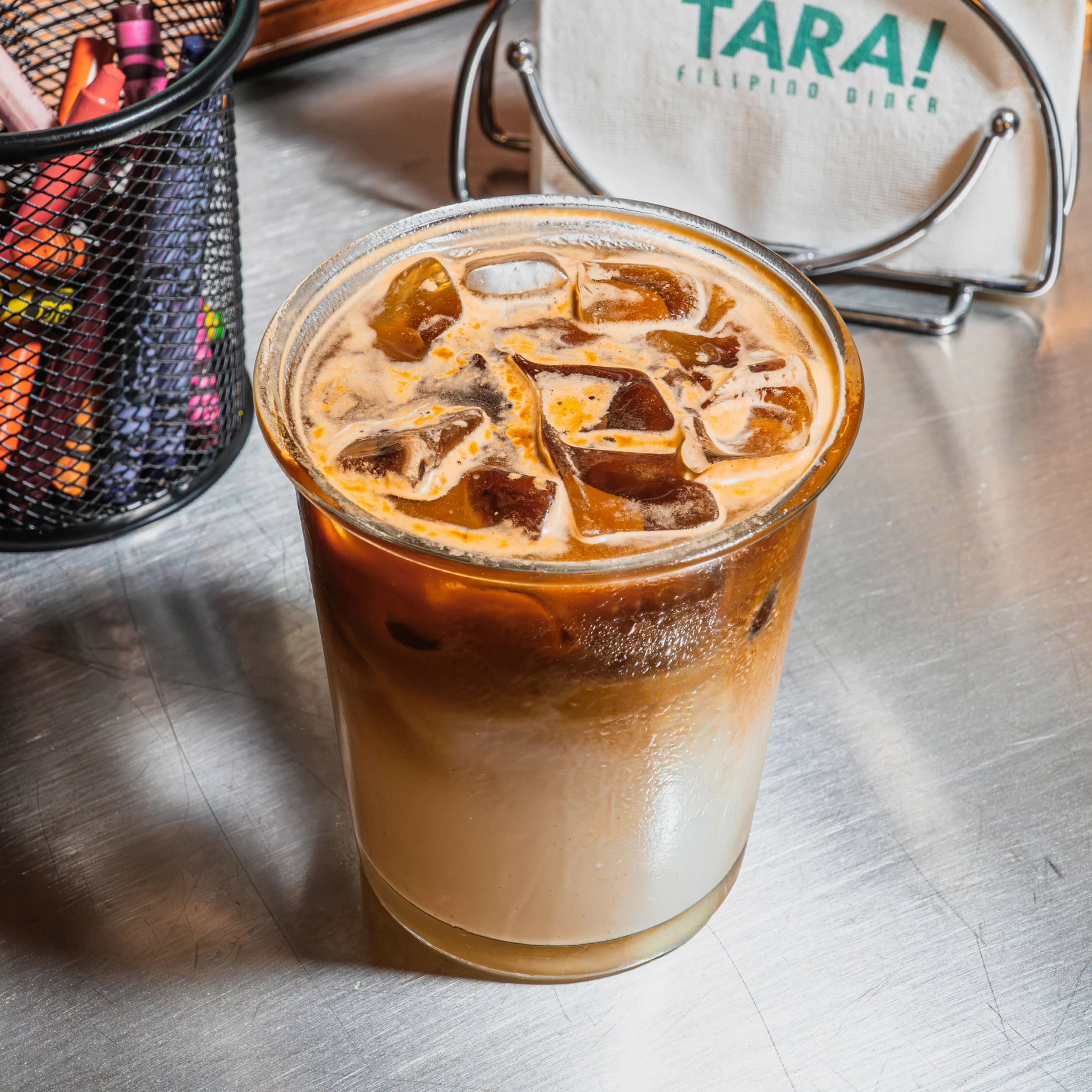 A layered iced coffee with milk and dark espresso sits on a metal café counter, topped with large ice cubes; warm, refreshing tones suggest a cozy café setting.