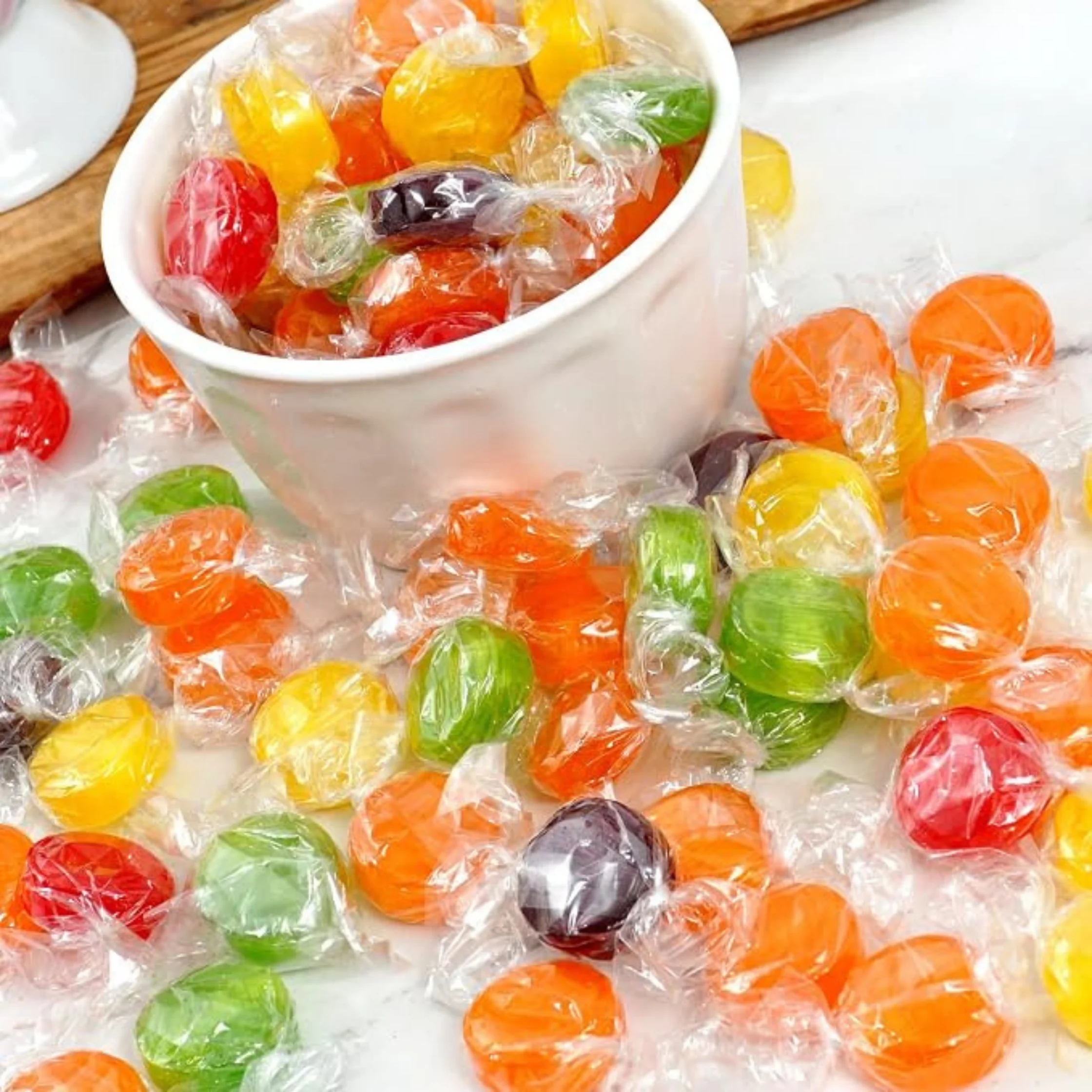 A white bowl brimming with individually wrapped, brightly colored hard candies spills across a tabletop, creating a cheerful, festive display.