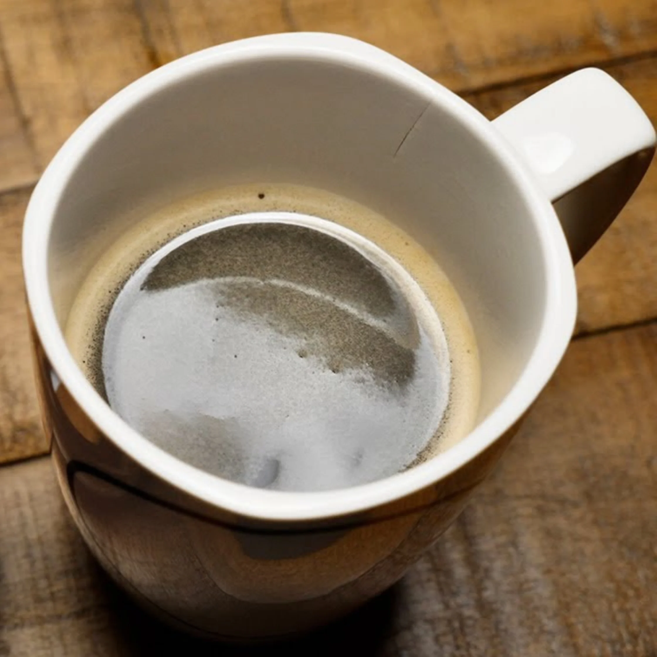 Overhead view of an empty white coffee mug with a ring of dried coffee residue and foam inside, resting on a rustic wooden table.