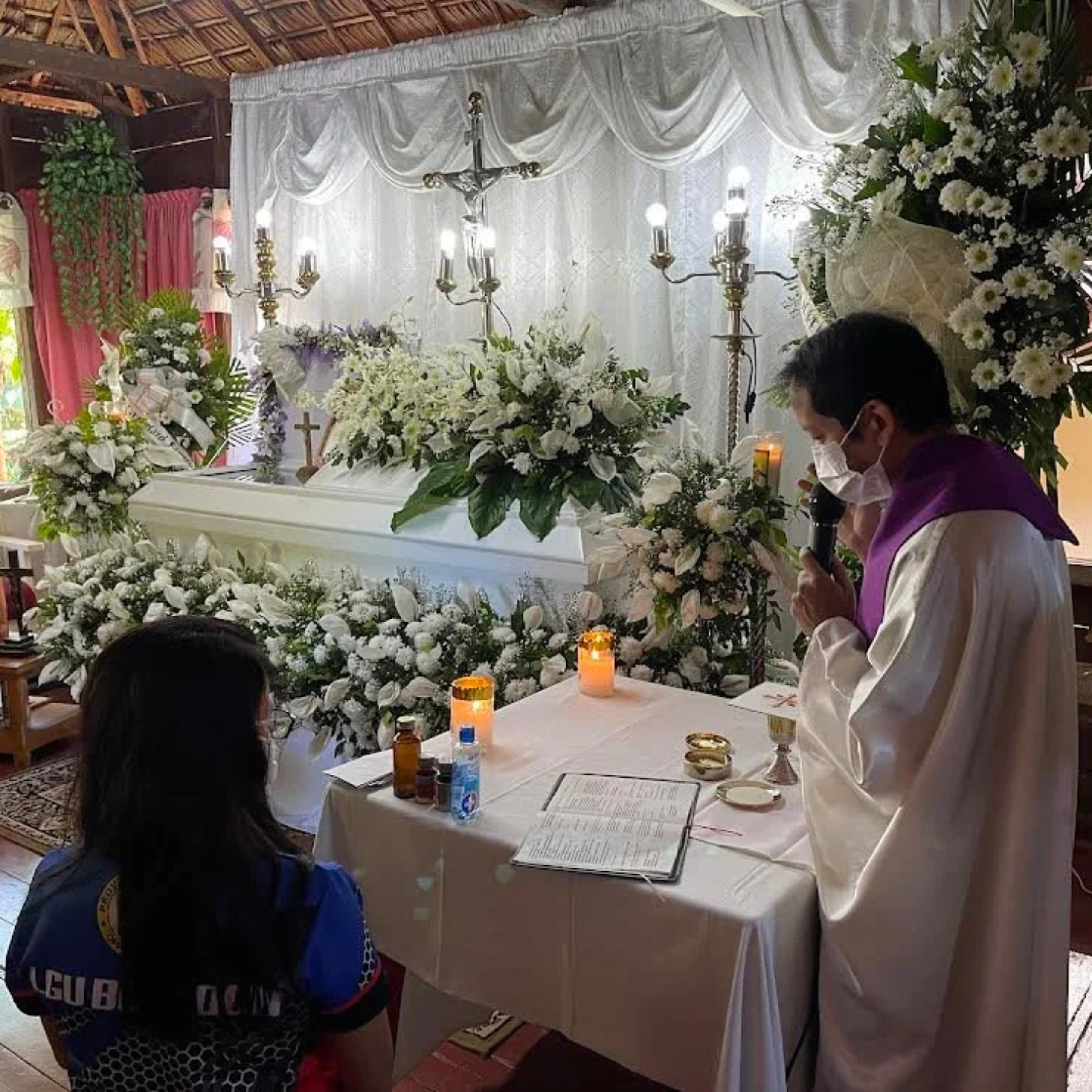 A masked priest prays beside a flower-covered white coffin as mourners gather in a candlelit funeral chapel, creating a solemn, reverent atmosphere.