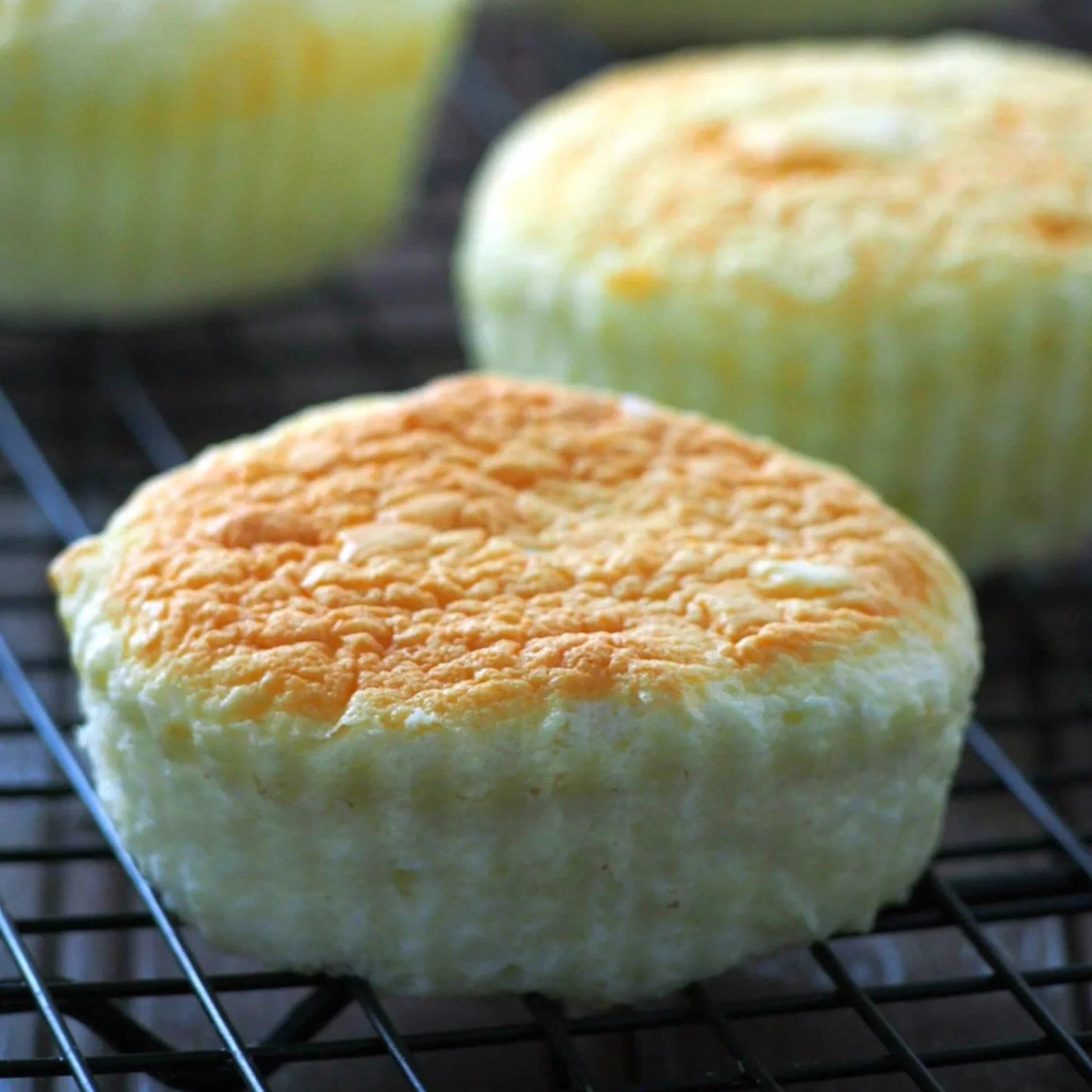 Golden-topped, fluffy baked cheese breads cool on a black wire rack, with two more softly blurred behind them; the scene feels warm and appetizing.