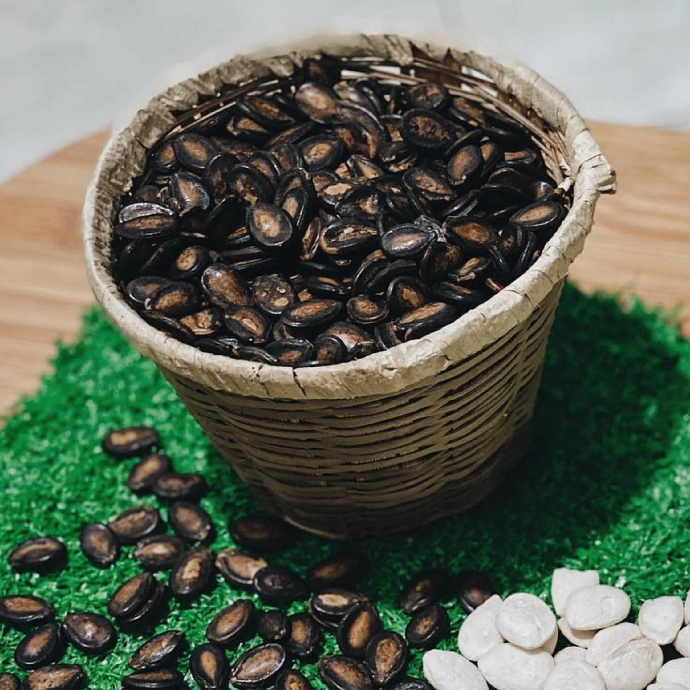 A rustic woven basket brimming with glossy dark seeds sits on a green mat, with more scattered nearby and pale pieces at the edge.