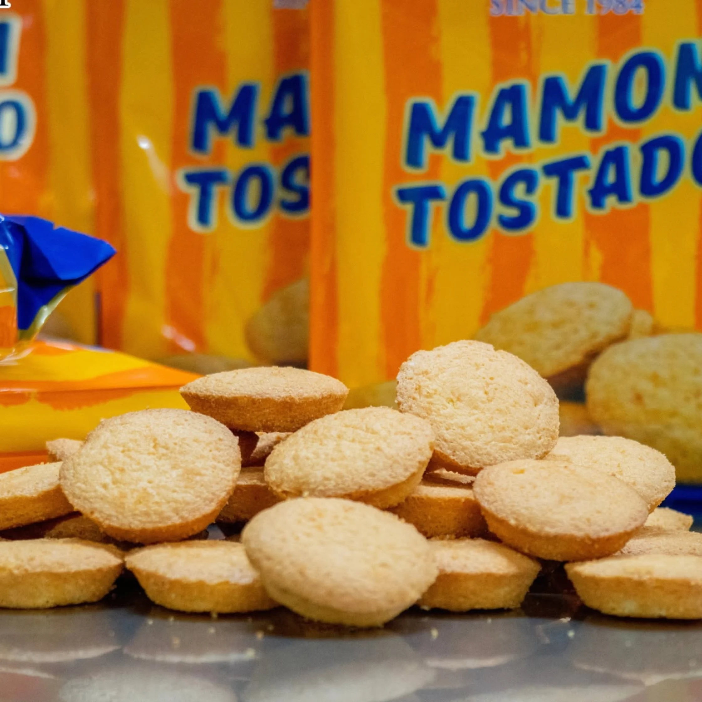 Golden toasted coconut biscuits piled in front of bright orange Mamon Tostado packages; a warm, cheerful close-up showcasing the crunchy snack.